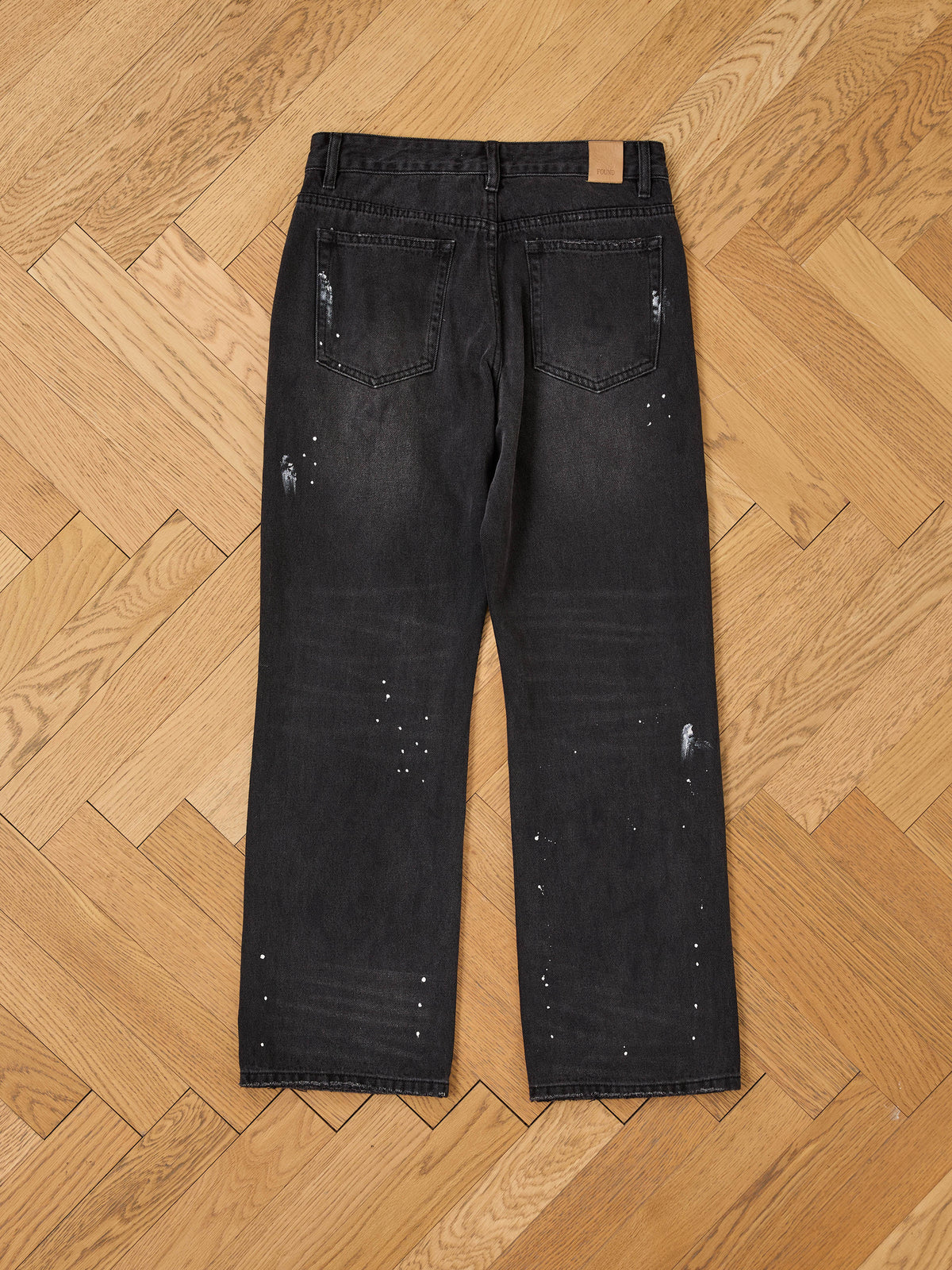 The Monet Paint Jeans by FOUND are vintage black denim with paint splatter and distressed details, shown flat from the back on a wooden herringbone floor.