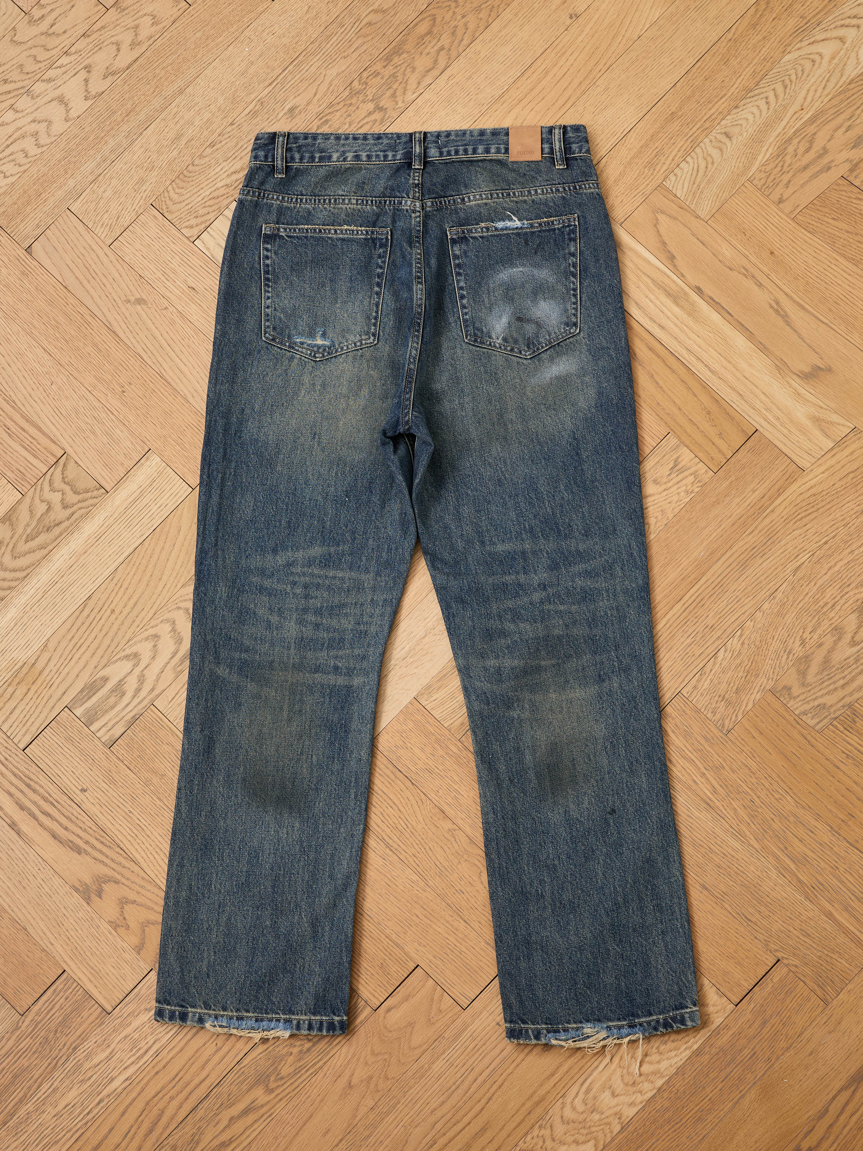 A pair of FOUND Monet Paint Jeans in vintage blue with worn patches and frayed hems, unisex relaxed fit, laid flat on a light wood herringbone floor and shown from the back.