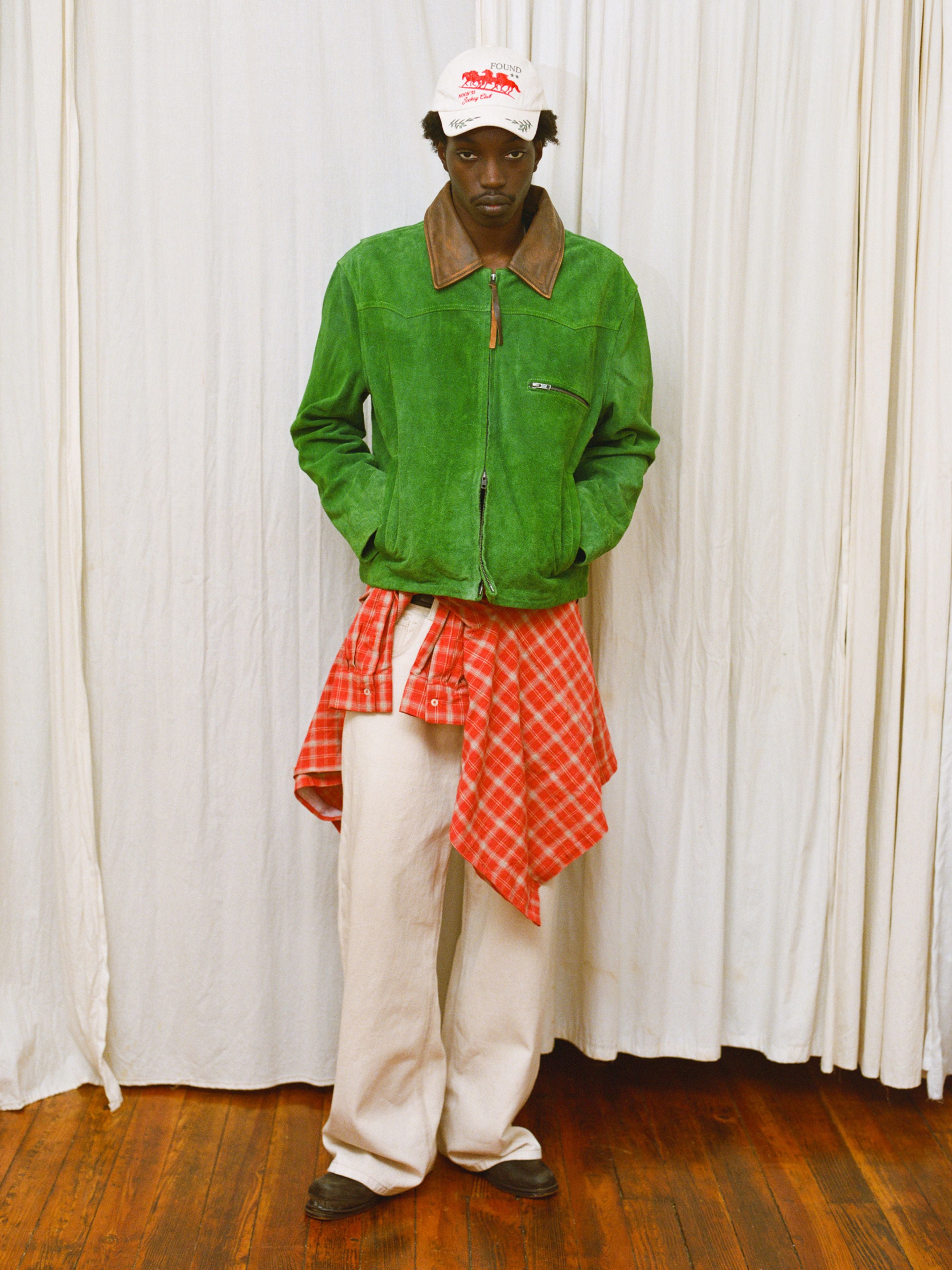 A person in a FOUND Suede Western Barn Jacket, white pants, brown shoes, a red plaid shirt tied at the waist, and a white cap stands in front of white curtains on a wooden floor.