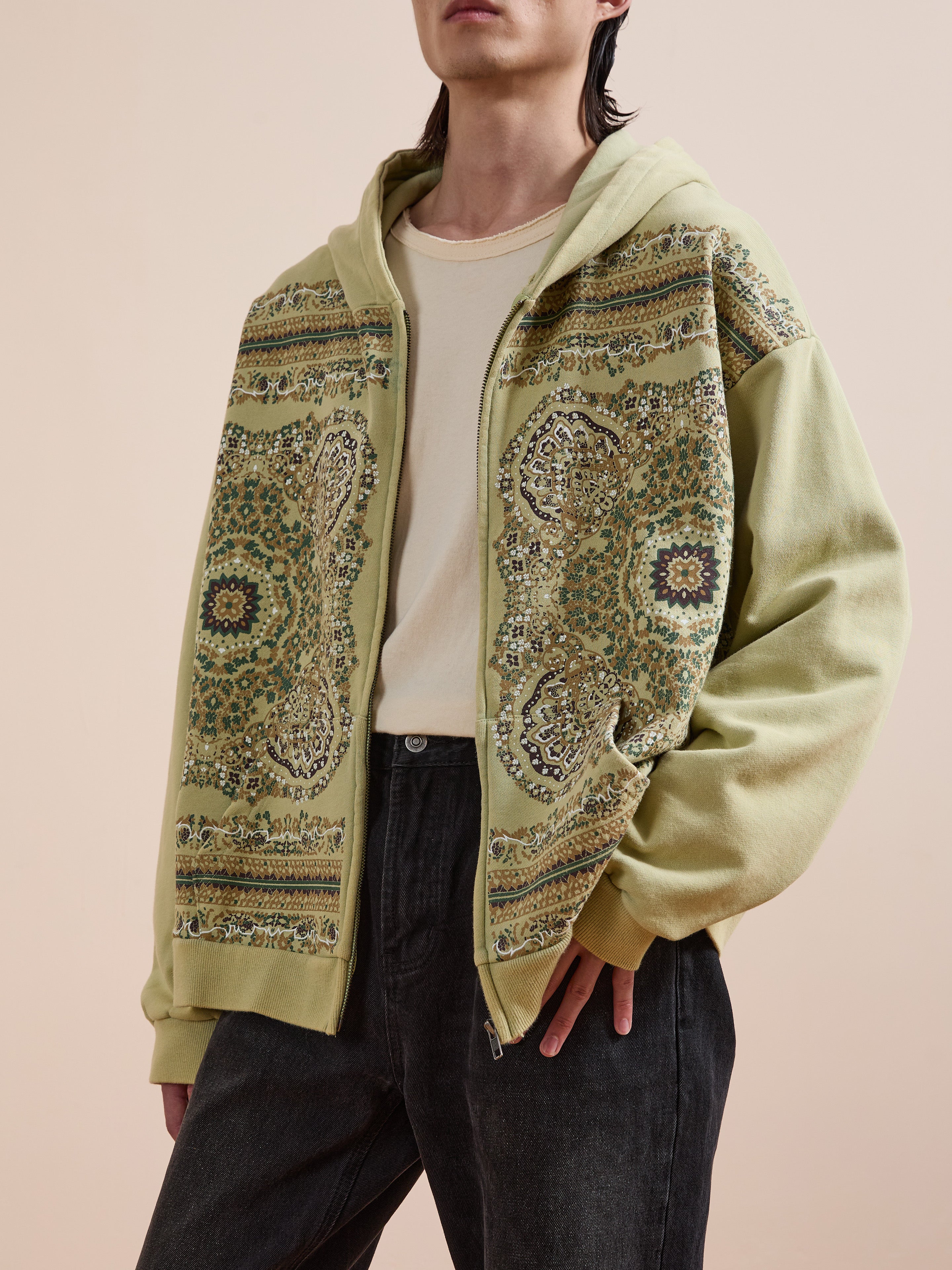 A person wears a light green FOUND Motif Zip-Up Hoodie over a cream t-shirt and black jeans, standing against a plain beige background—a must-have streetwear essential.