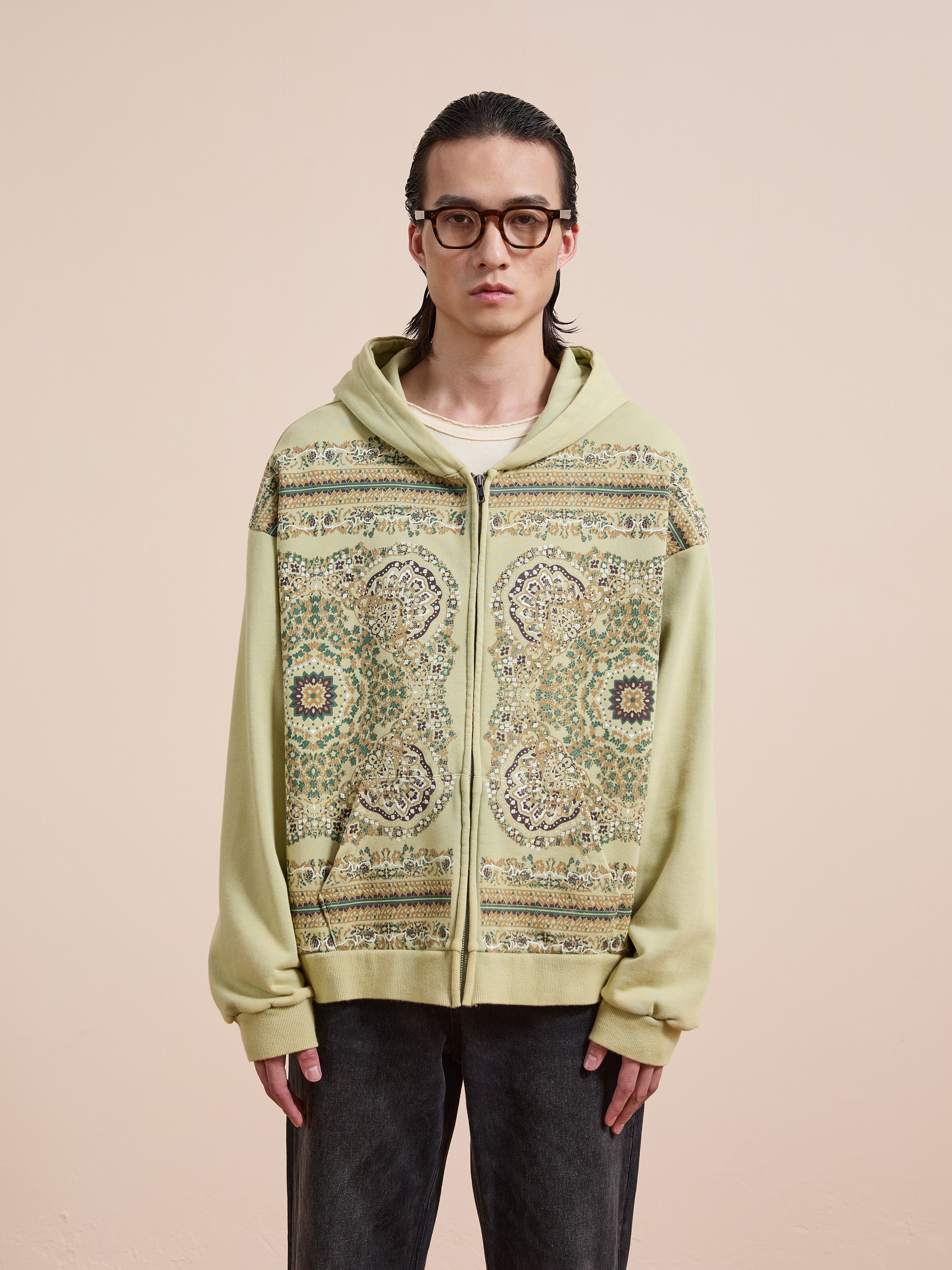 A person with long dark hair and glasses wears the FOUND Motif Zip-Up Hoodie in light green over a white shirt, standing against a beige background—a must-have for streetwear fans.