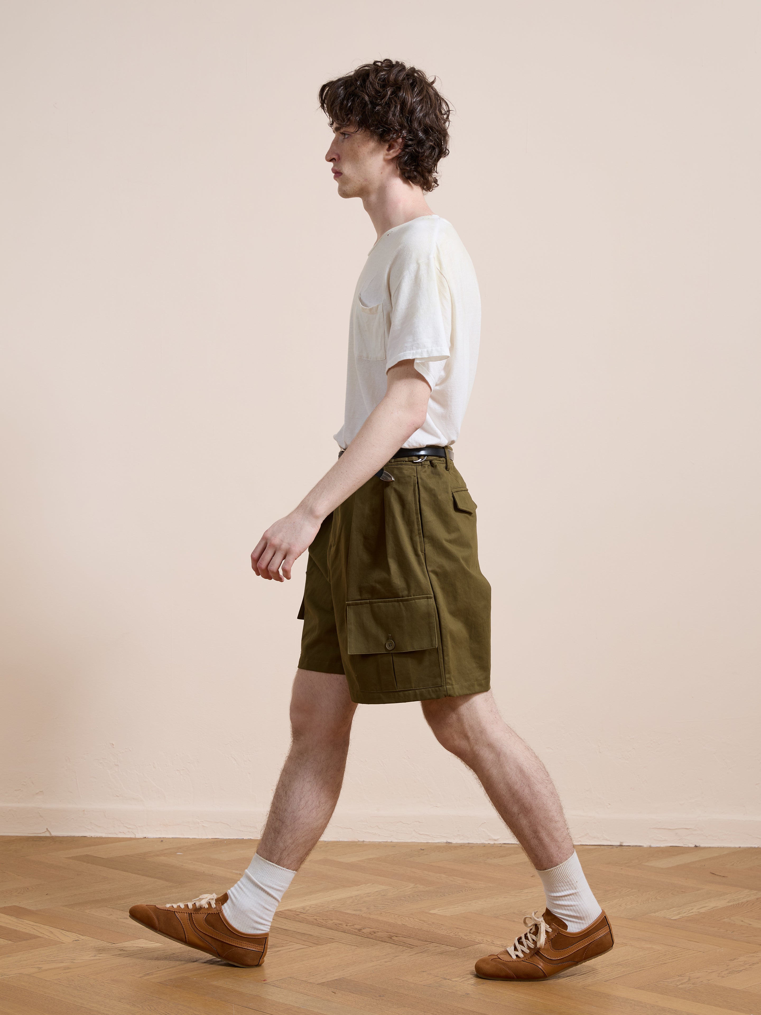 A man in a white t-shirt, Found Multi Pocket Shorts, white socks, and brown shoes walks indoors on a wooden floor against a beige wall. The Found shorts add practical style to his casual outfit.