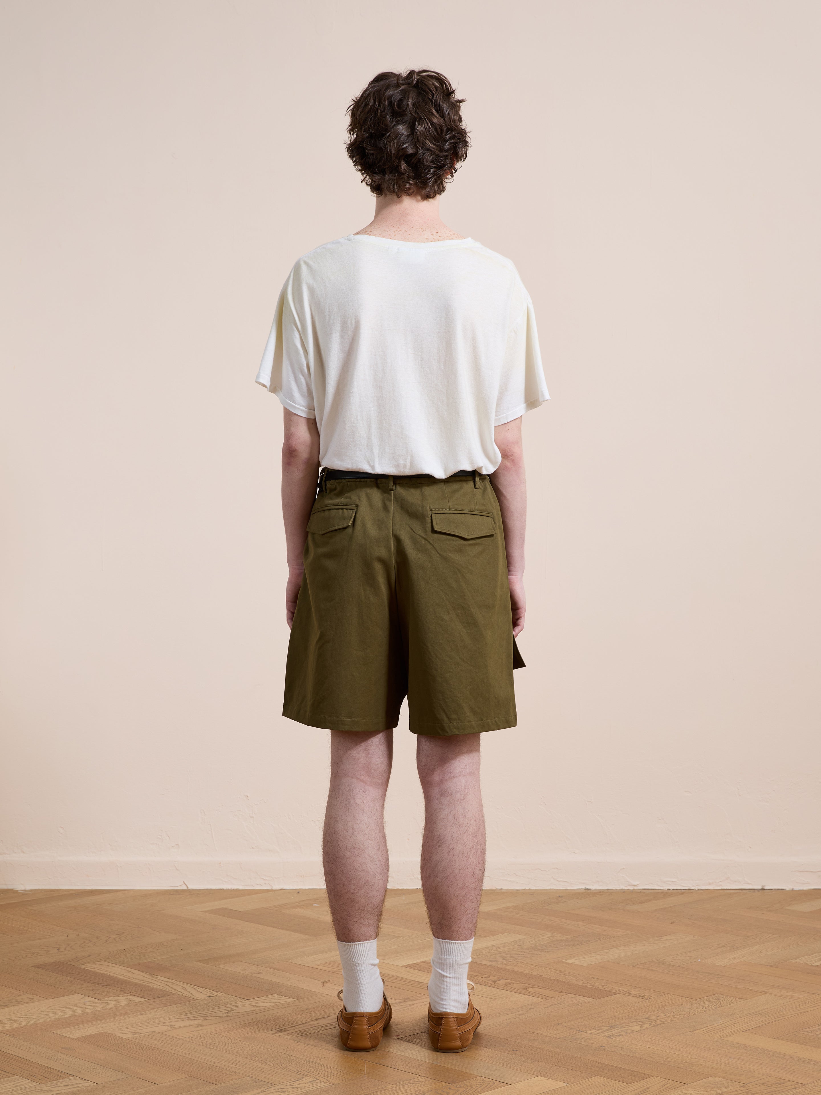 A person stands indoors on a wooden floor, facing away from the camera, wearing a white t-shirt, white socks, brown shoes, and Found Multi Pocket Shorts in olive green.