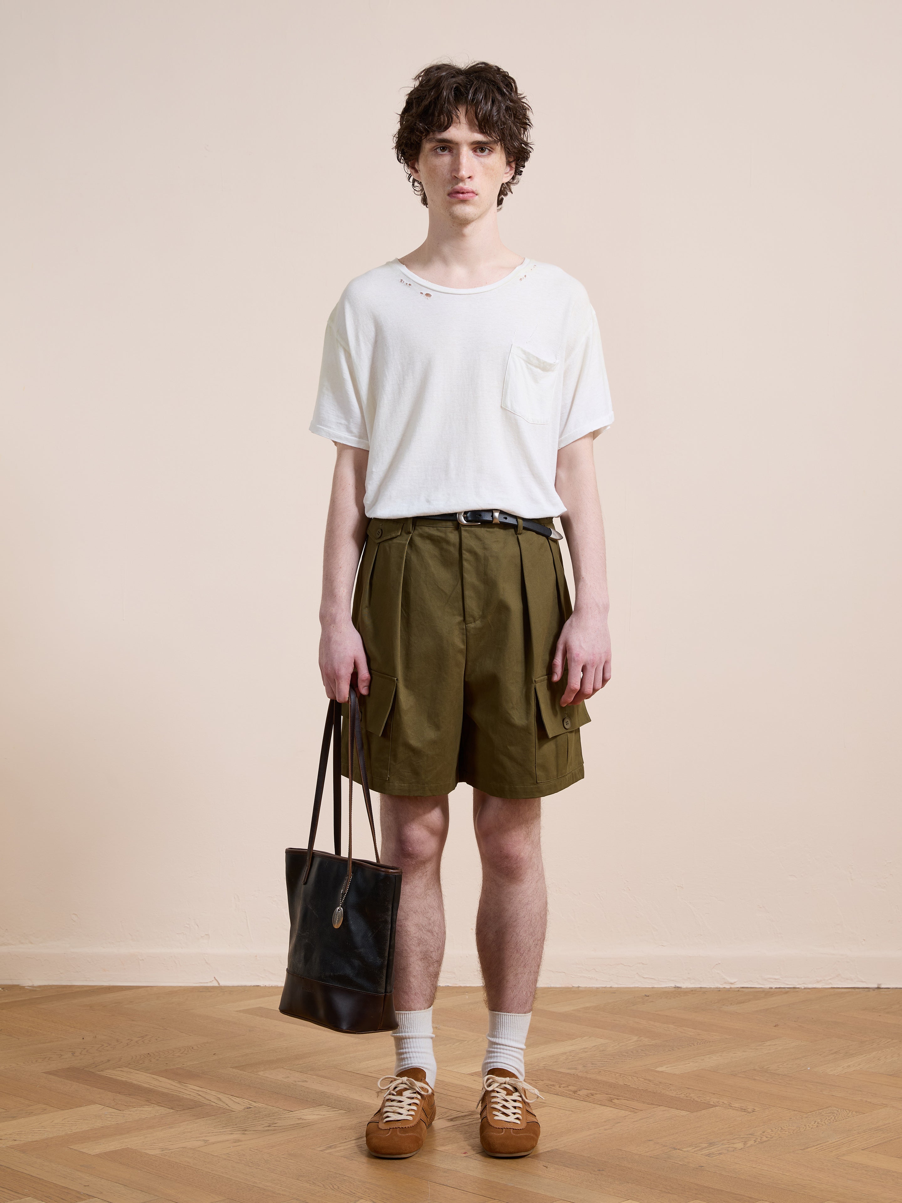 A young person stands against a plain background wearing Found Multi Pocket Shorts in olive green, a white t-shirt, brown shoes, and holds a black tote bag in their left hand.
