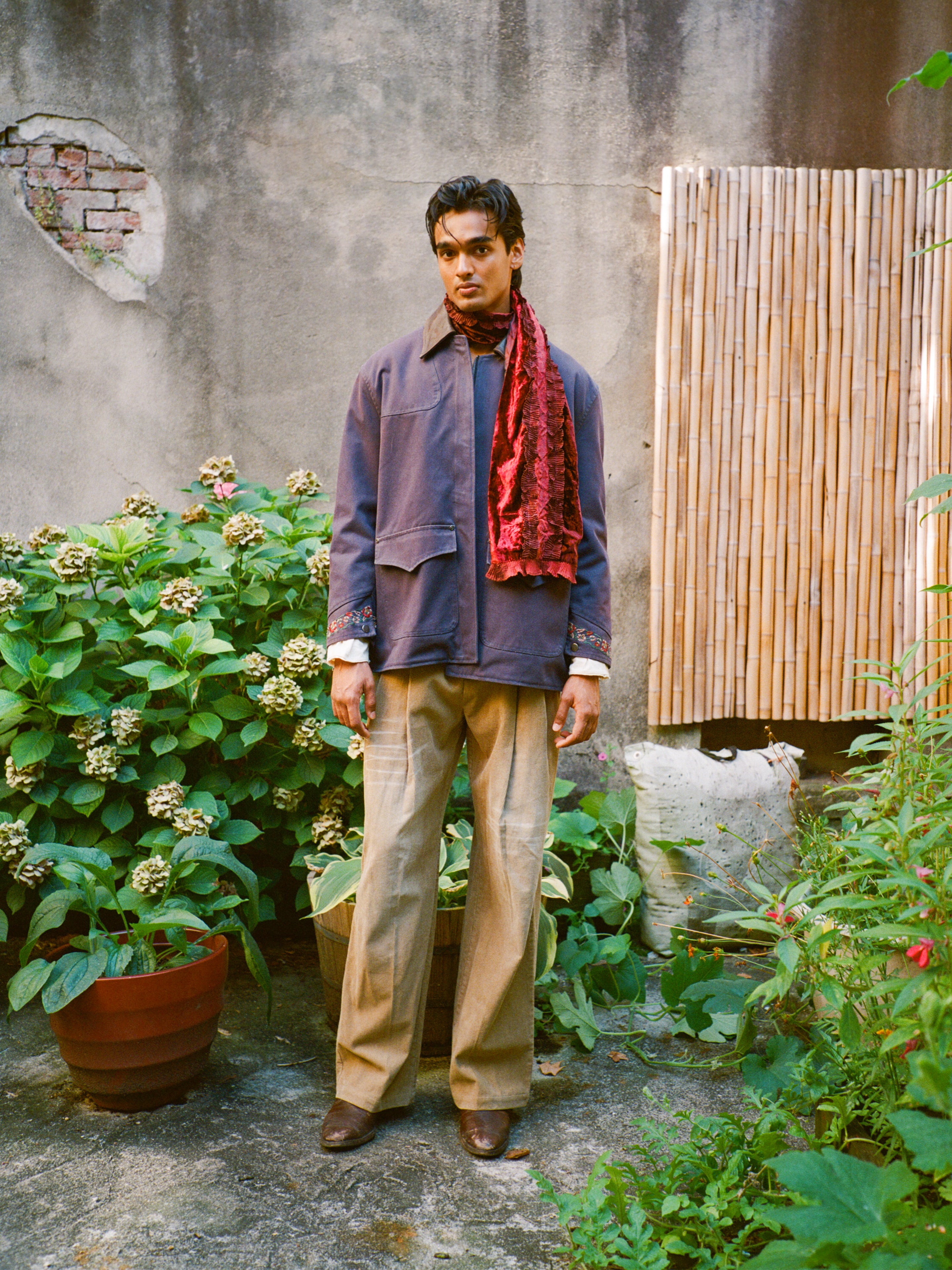 A man stands in a garden wearing the Found Canvas Distressed Barn Jacket, paired with tan pants and a red scarf. Potted plants, hydrangeas, and a bamboo screen against a weathered wall set a tranquil scene.