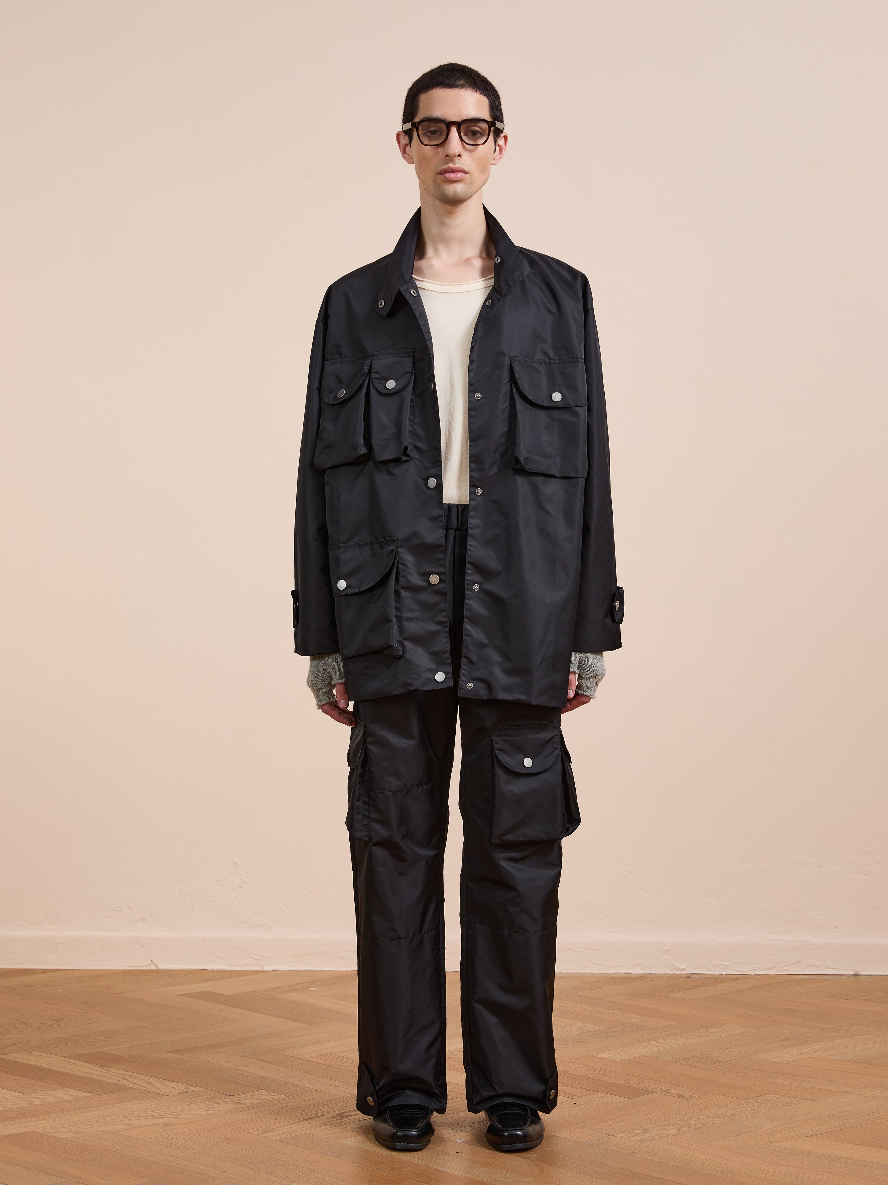 A person faces the camera indoors against a beige background, wearing FOUND's Cargo Nylon Jacket in black over a white shirt, paired with black cargo pants and glasses.