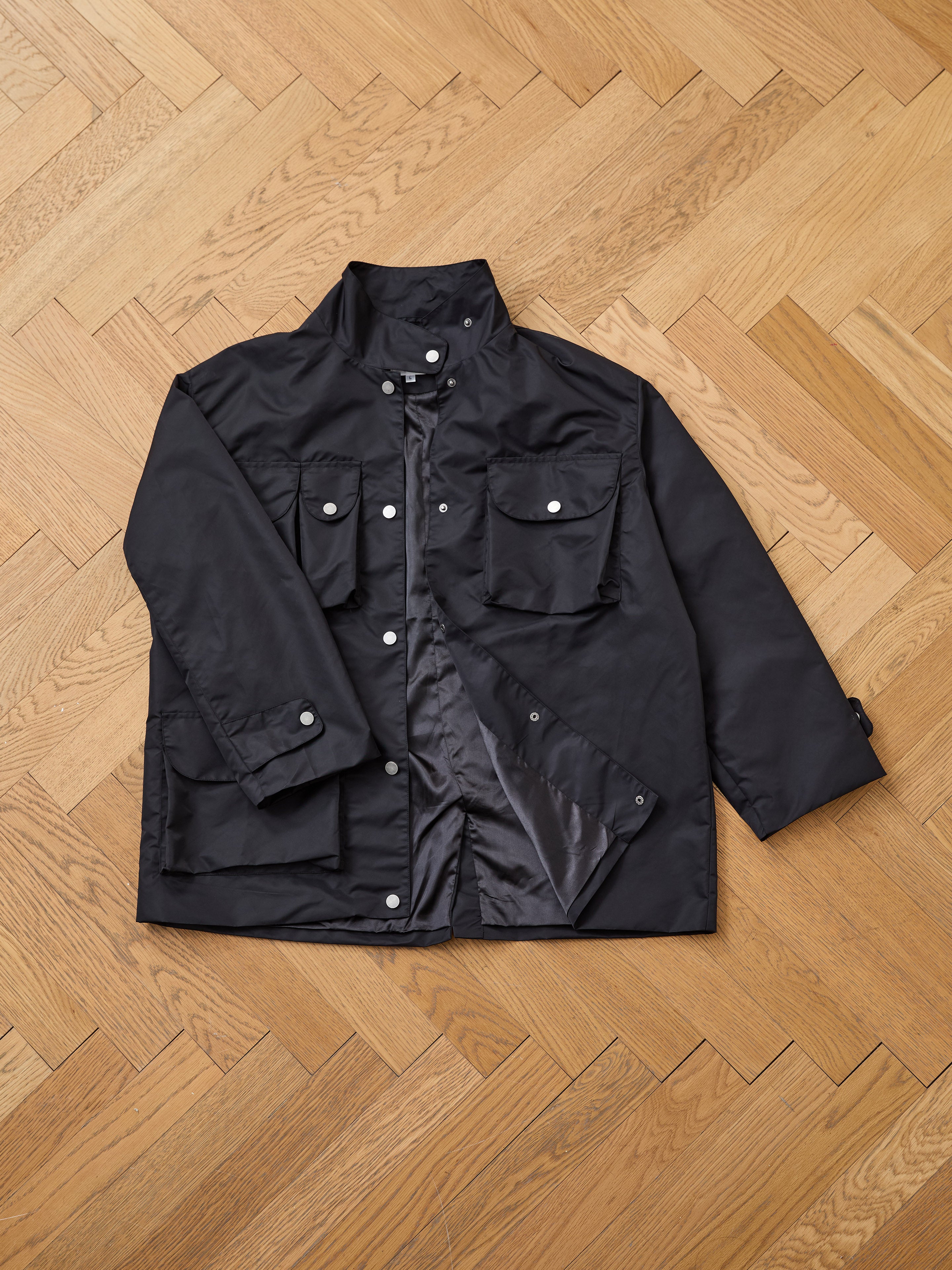The FOUND Cargo Nylon Jacket is a black unisex, long-sleeve jacket with multiple front pockets and snap buttons, shown laid flat on a wooden parquet floor.