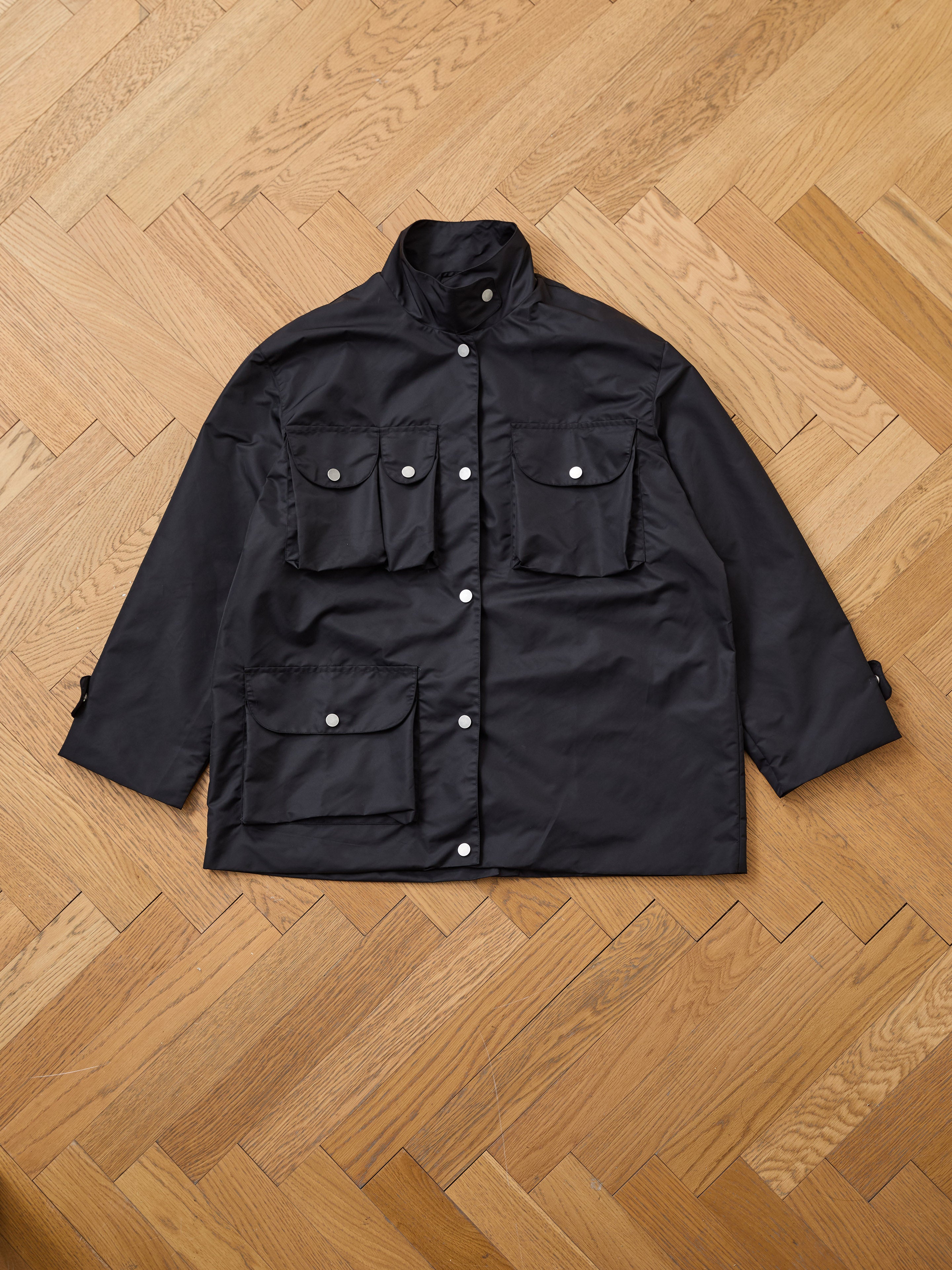 The FOUND Cargo Nylon Jacket, a black unisex style with four front flap pockets and silver buttons, is displayed flat on a light wood herringbone floor.