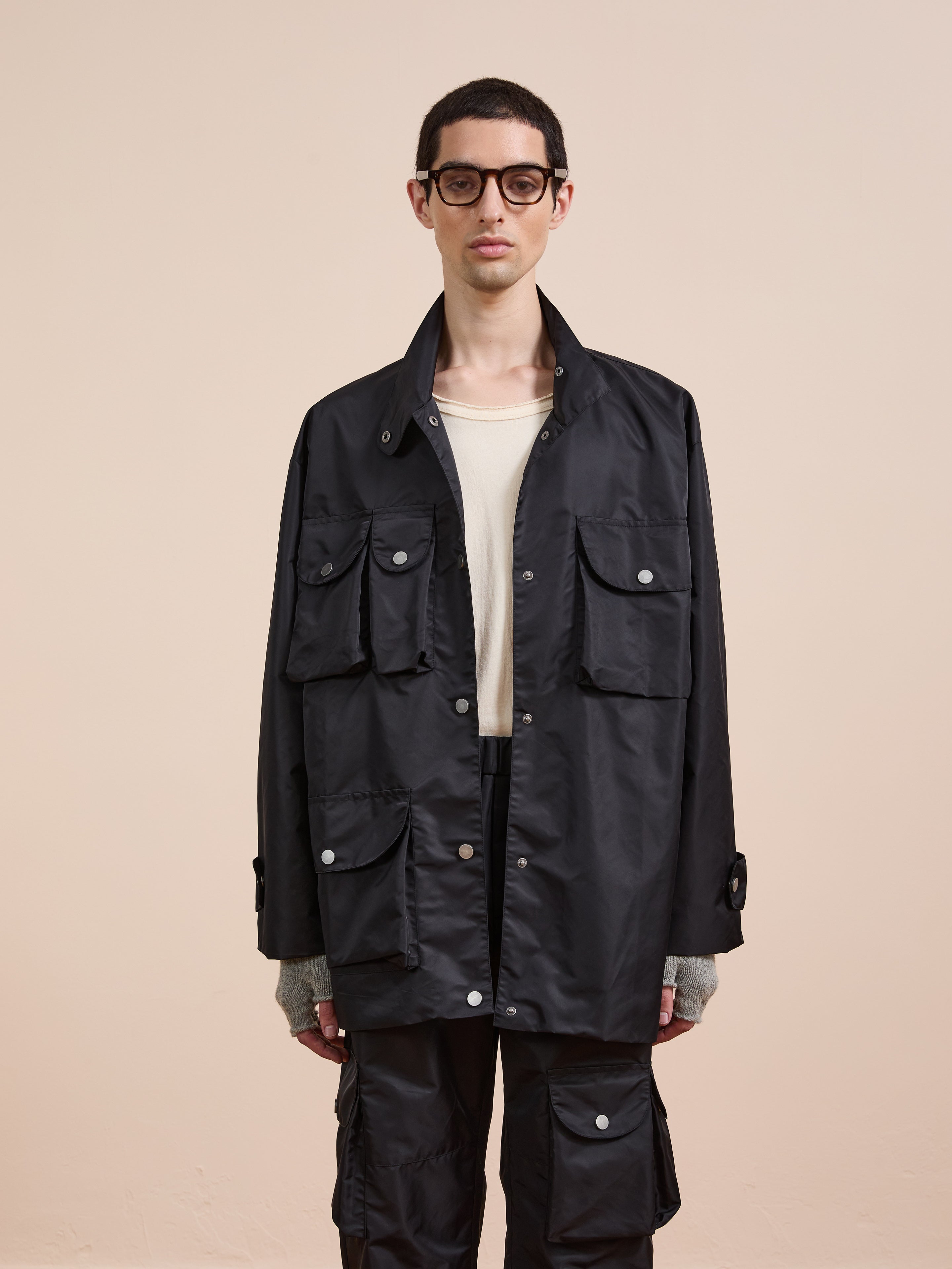 A person wears the FOUND Cargo Nylon Jacket in black, paired with dark pants, a cream shirt, and glasses, standing against a plain beige background.