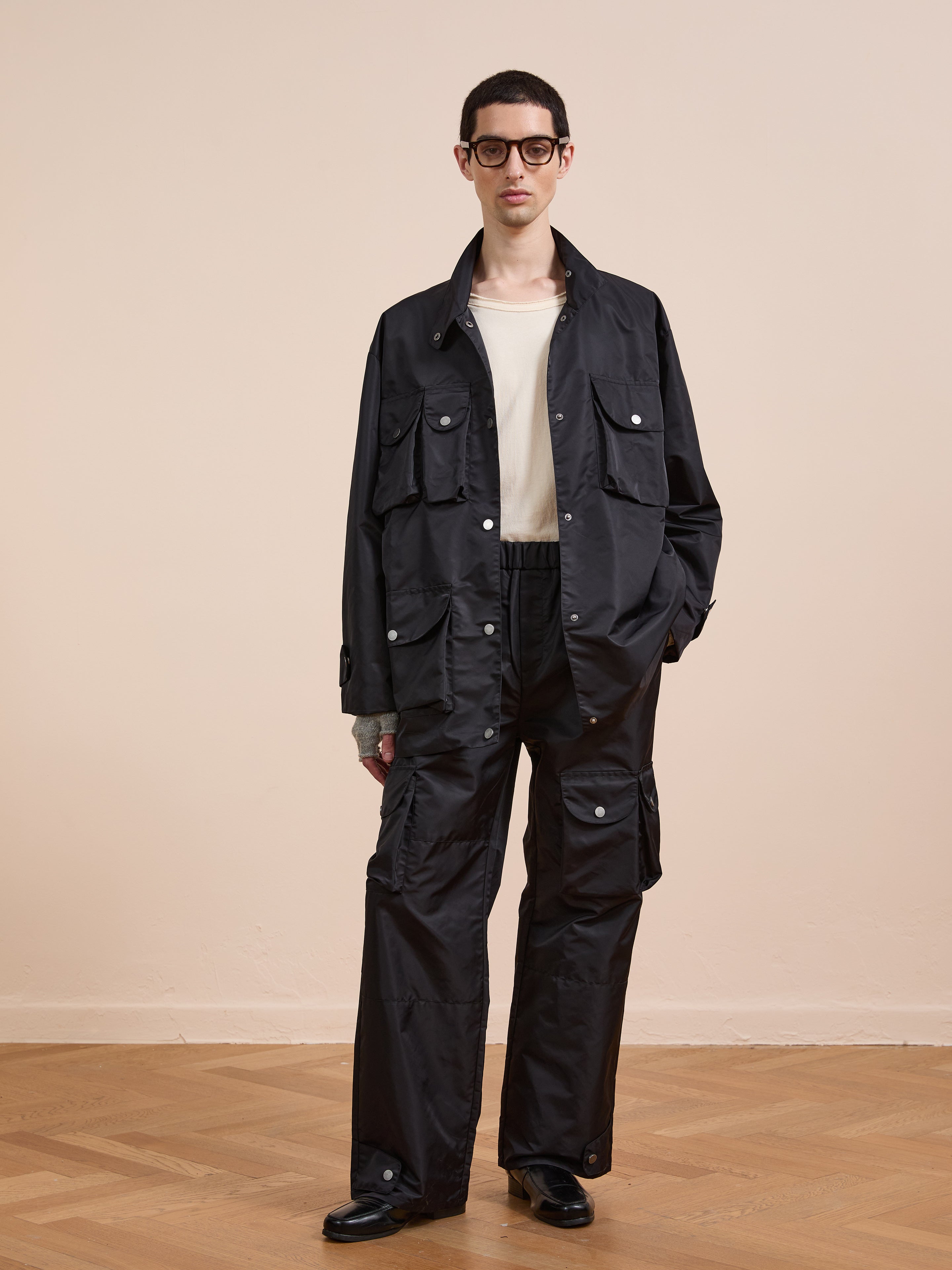 A person wears the FOUND Cargo Nylon Jacket styled with matching utility pants, a white shirt, dark shoes, and glasses, standing on a wooden floor against a plain beige wall.