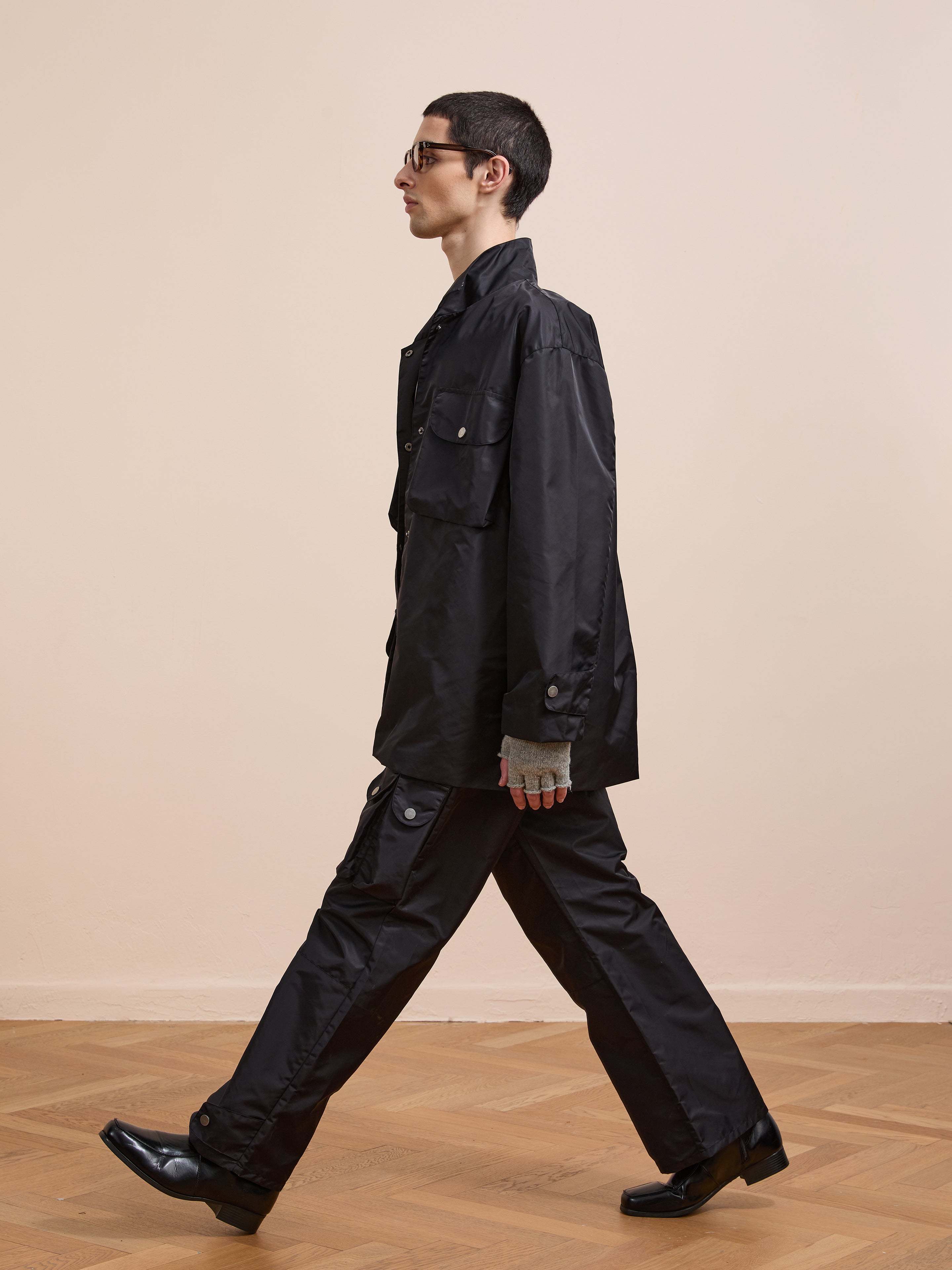 A person with short dark hair and glasses wears the FOUND Cargo Nylon Jacket, black pants, and black shoes while walking indoors on a wooden floor against a plain beige wall.