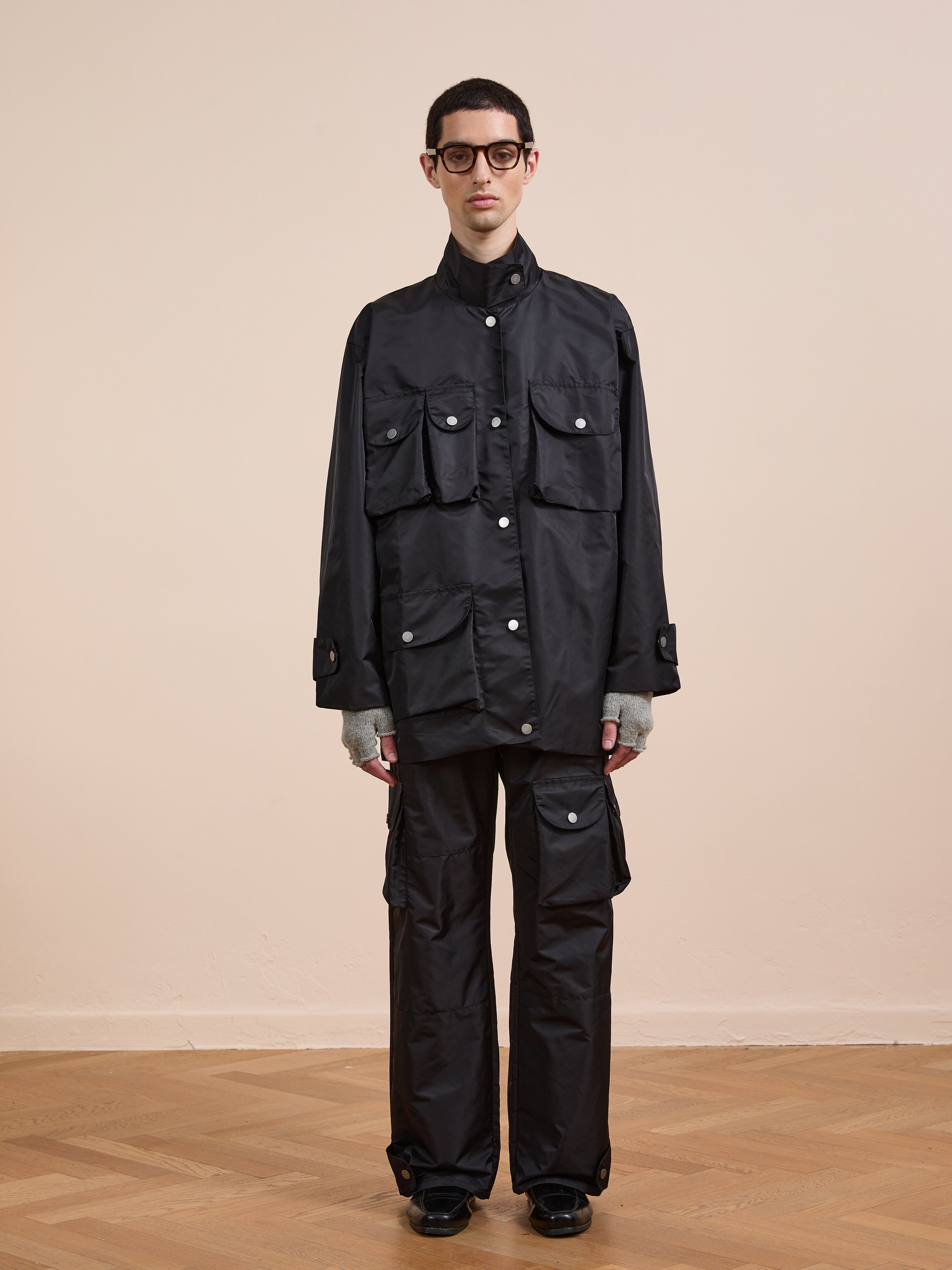 Indoors on a wooden floor, someone wears black glasses, grey gloves, black cargo pants, and the FOUND Cargo Nylon Jacket—a unisex black utility coat featuring multiple front pockets.