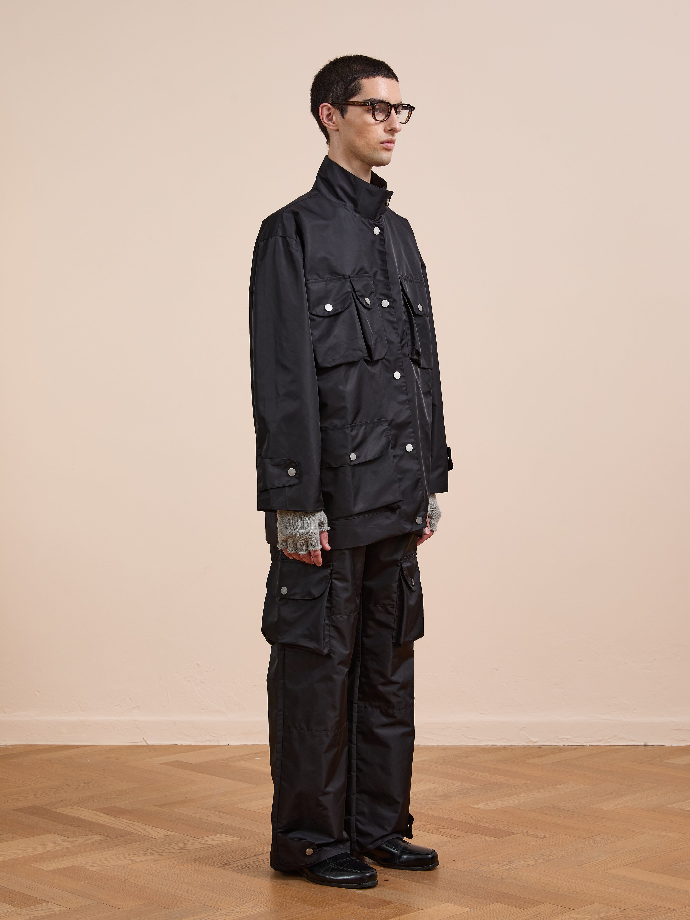 A person stands indoors on a wooden floor against a beige wall, wearing the FOUND Cargo Nylon Jacket with matching cargo pants, gray gloves, black shoes, and glasses.