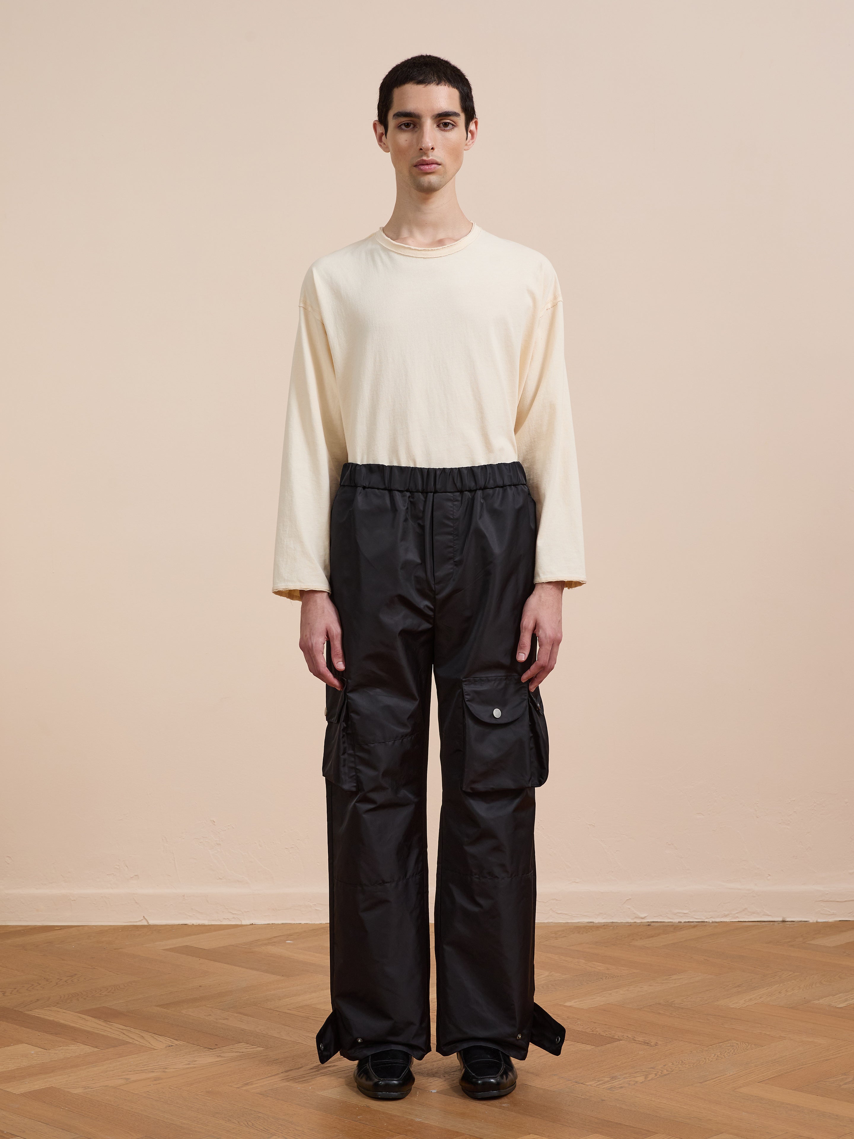A person stands indoors on a wooden floor wearing FOUND Nylon Cargo Pants—unisex black cargo pants with large pockets and a relaxed fit—paired with a cream long-sleeve shirt and black shoes, against a beige wall.
