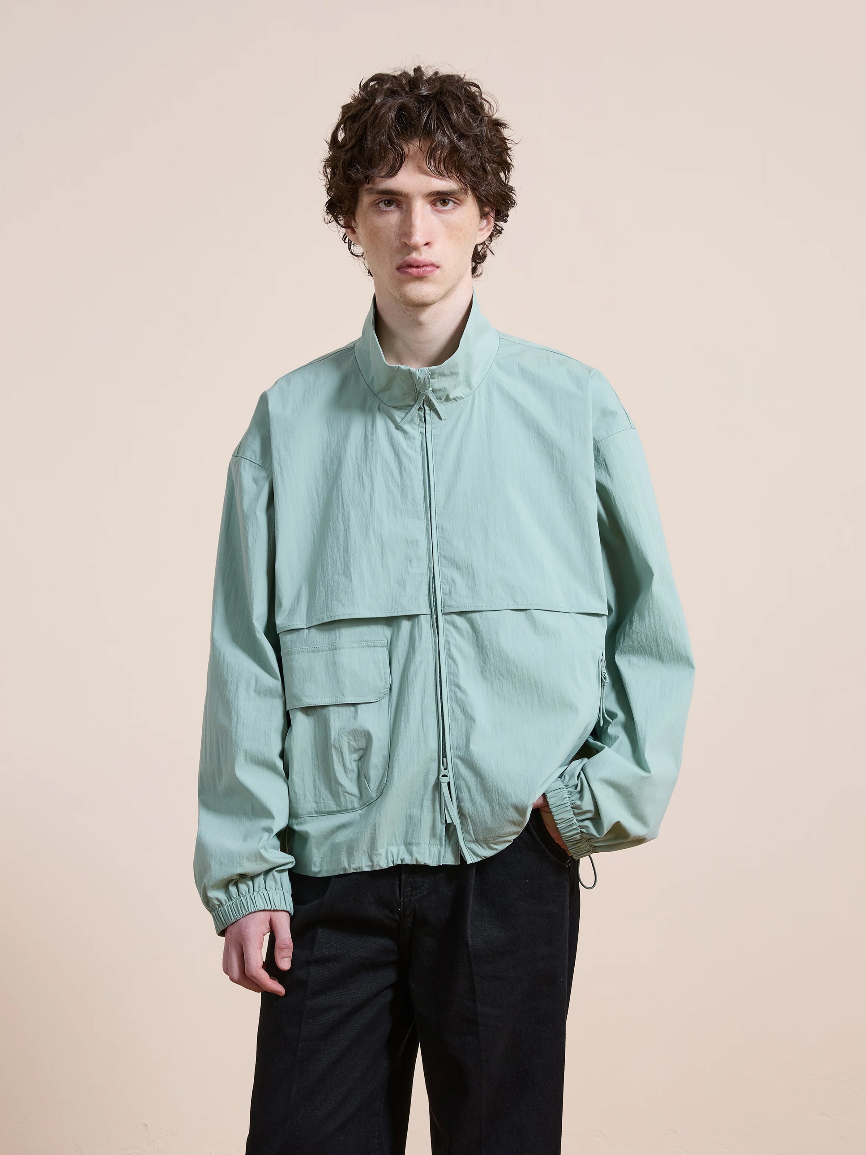 A person with curly hair wears the FOUND Nylon Pocket Track Jacket in light blue and black pants, standing against a plain beige background.