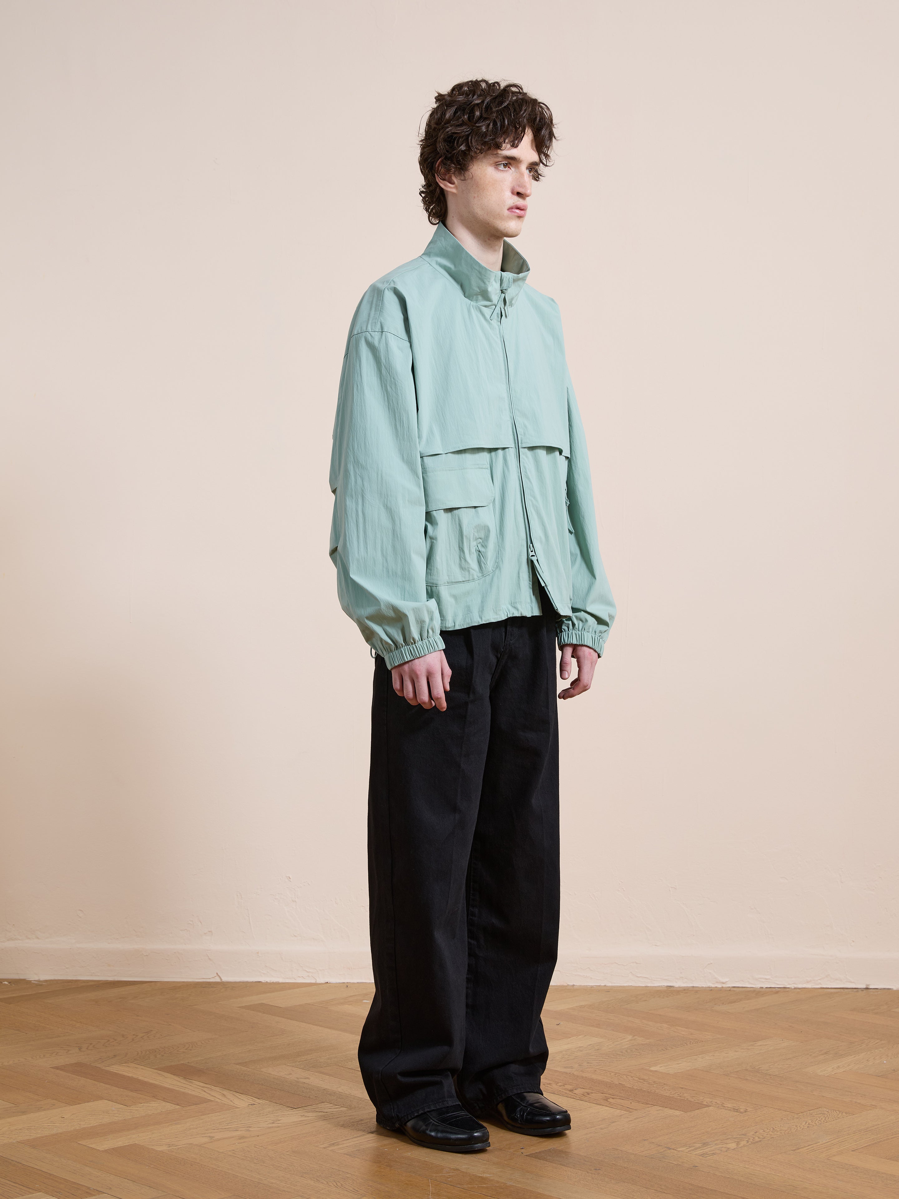 A person stands indoors in a FOUND Nylon Pocket Track Jacket, paired with black pants and black shoes, against a plain beige wall and wooden floor.