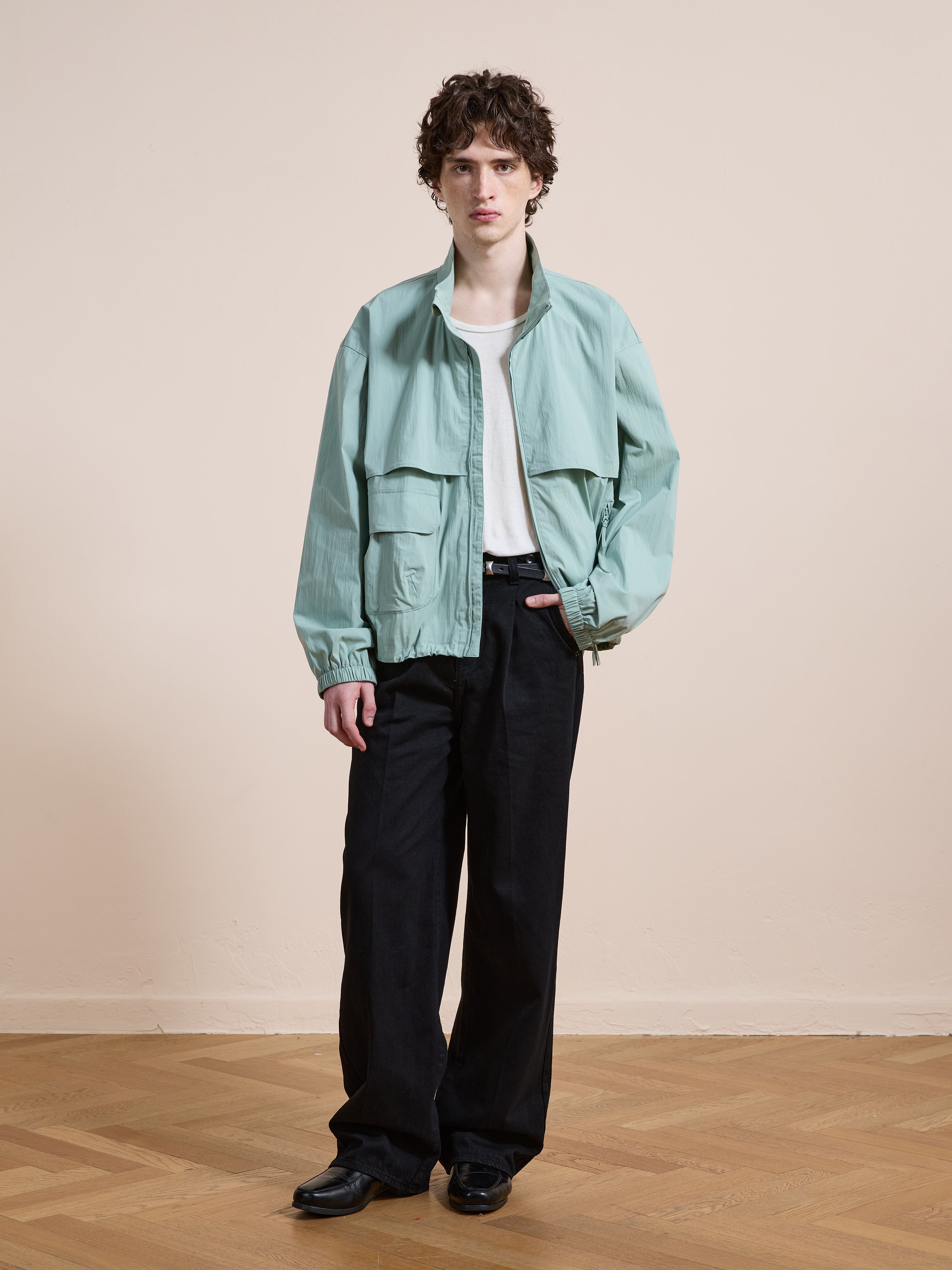 Indoors, a person wears the FOUND Nylon Pocket Track Jacket in light green with a white tee, black wide-leg pants, and black shoes, standing on a wooden floor against a plain beige wall.
