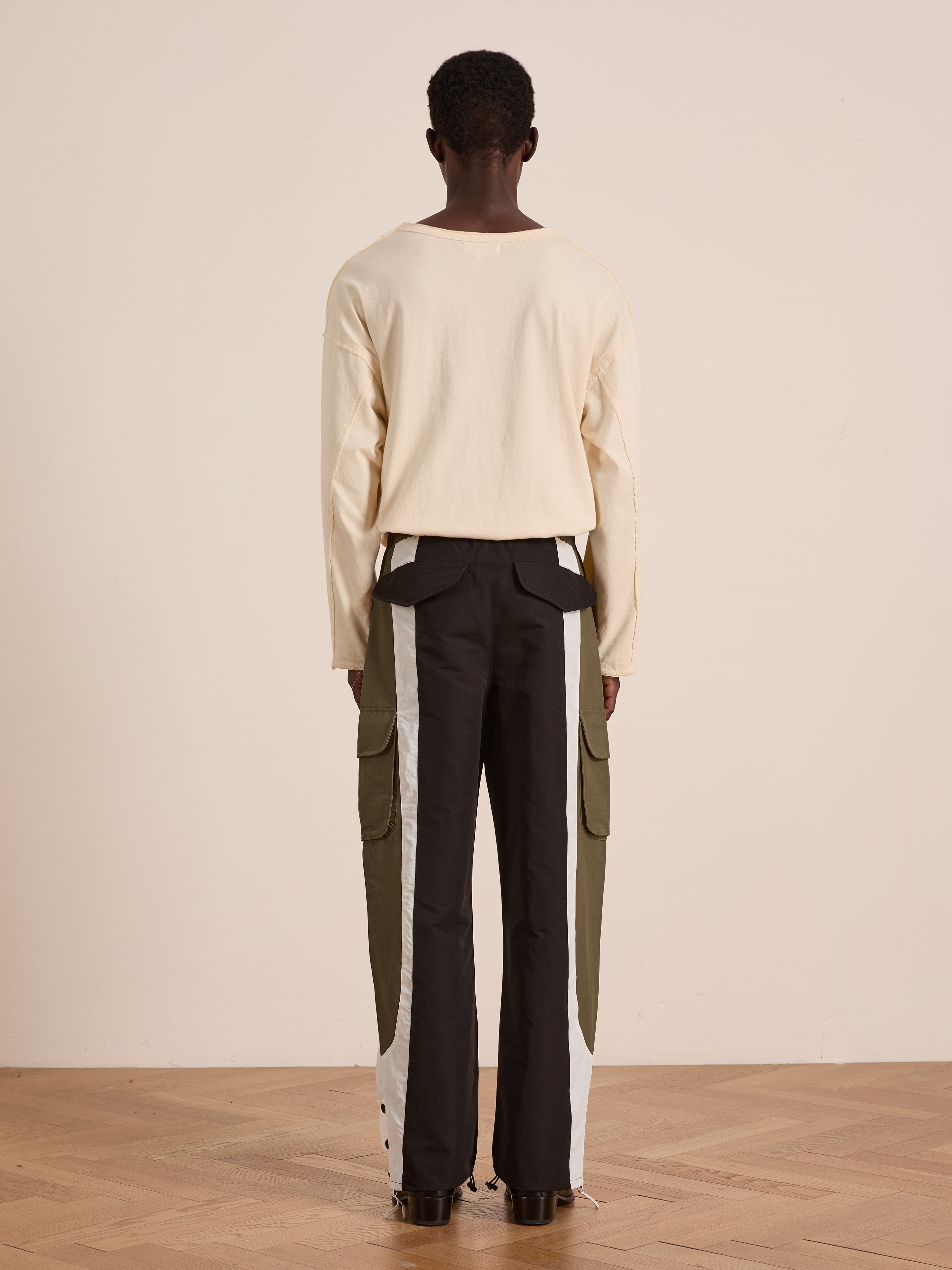 Standing indoors facing away, person wears a long-sleeve beige shirt with Found Sporting Club Track Pants—black with gray and olive green side panels and adjustable waistband. A versatile unisex choice for sportswear enthusiasts.