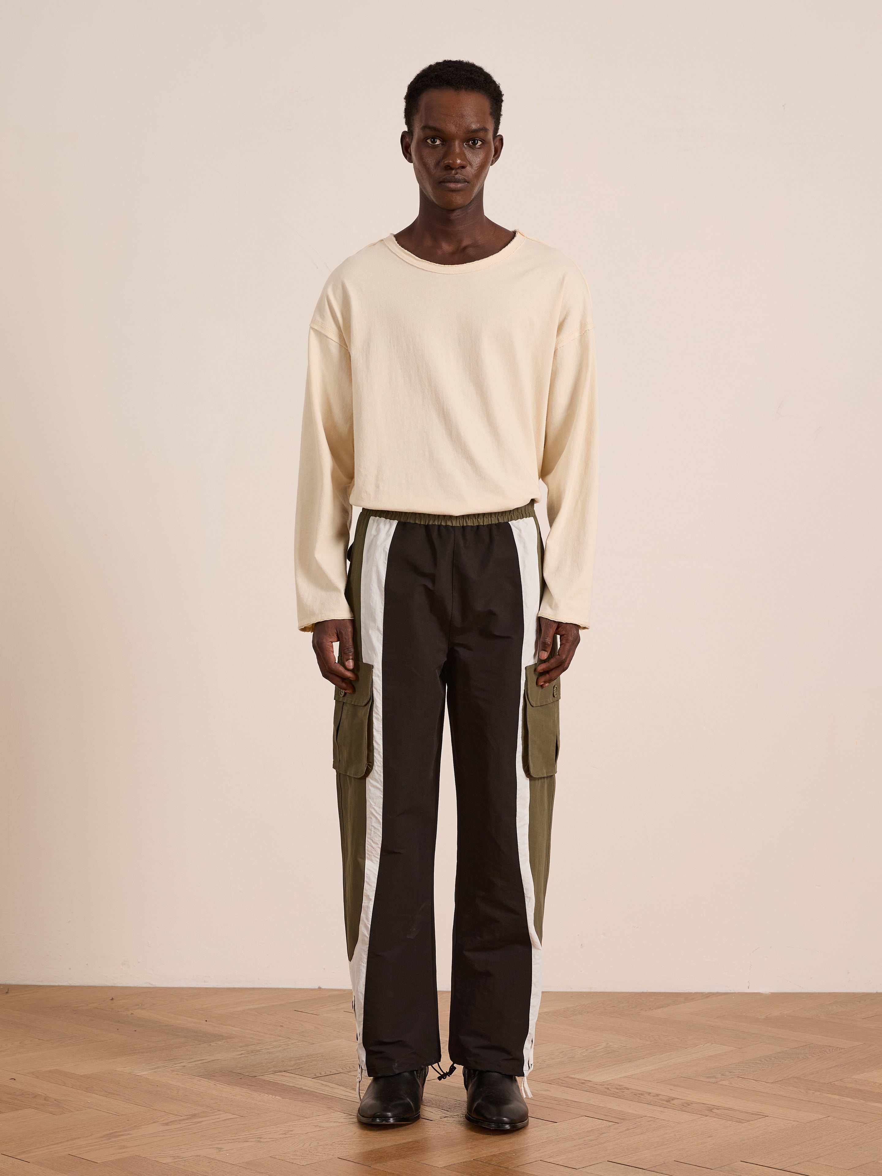 A person stands indoors wearing Found Sporting Club Track Pants—black with white and olive side panels and an adjustable waistband—styled as modern unisex sportswear against a plain background.