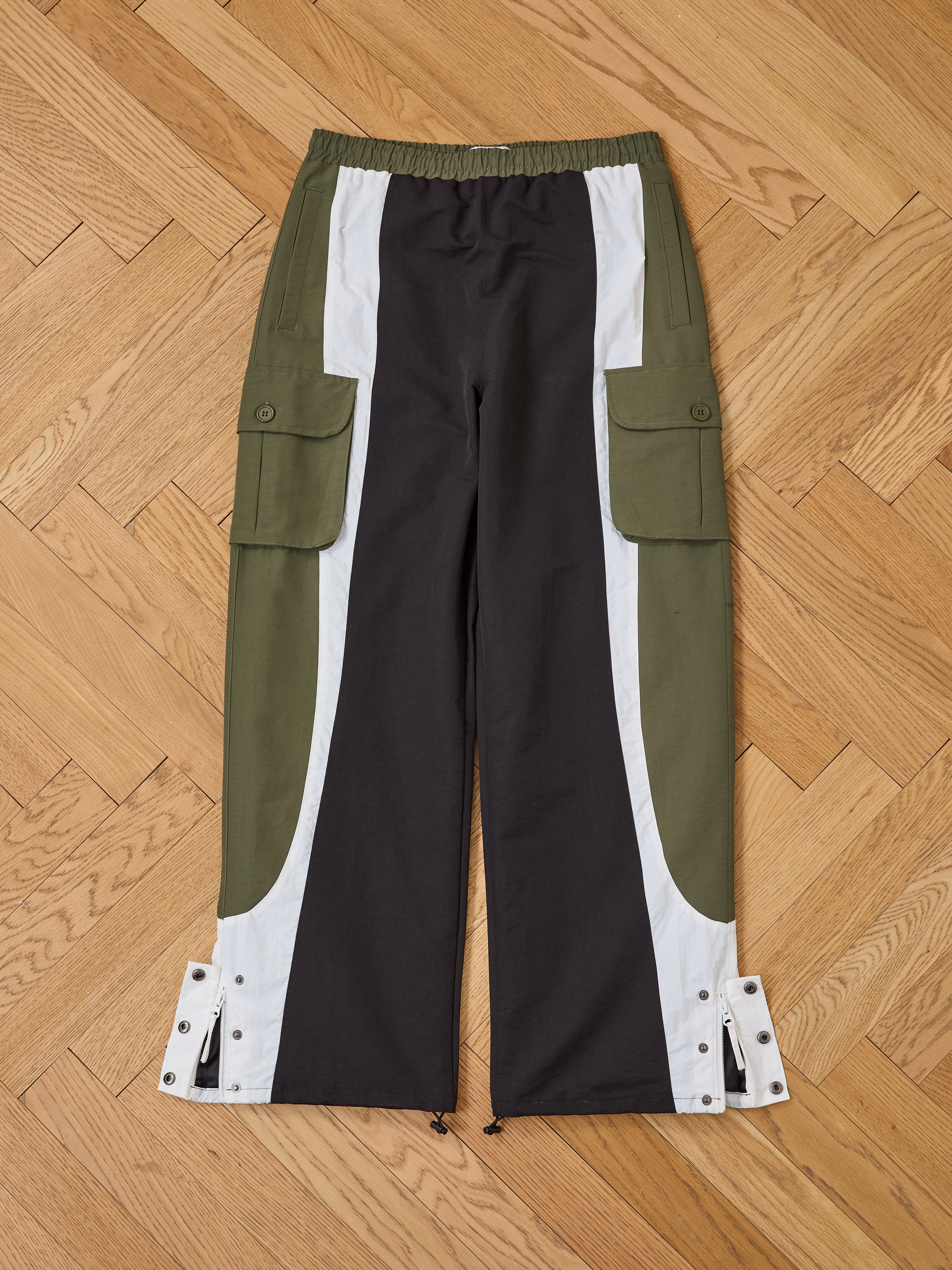 The Found Sporting Club Track Pants, a unisex sportswear piece with black, olive green, and white panels, side pockets, ankle snaps, and an adjustable waistband, is shown laid flat on a wooden floor.