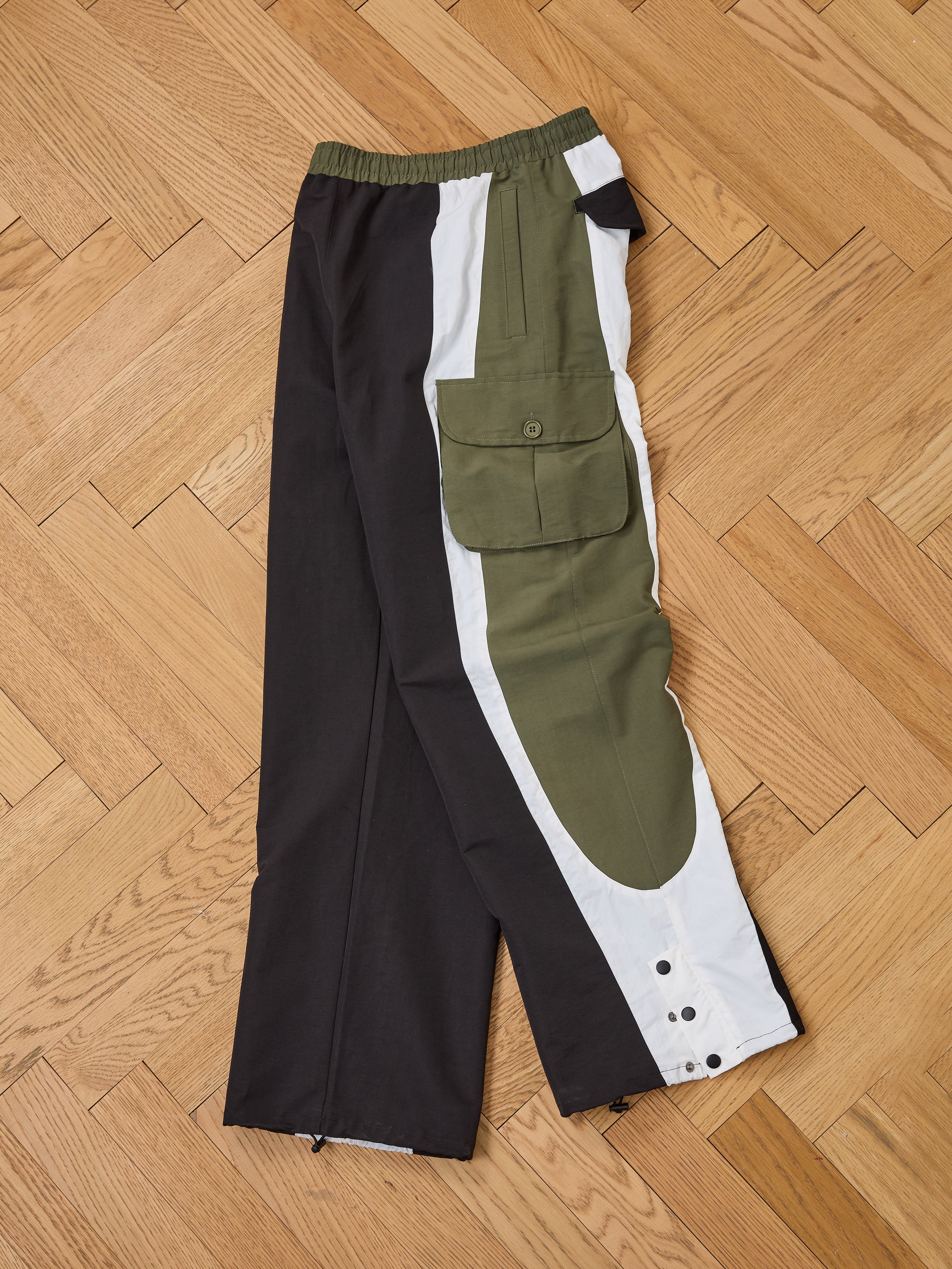 The Sporting Club Track Pants by Found are unisex color-block cargo pants in black, white, and olive green, with an adjustable waistband, large side pockets, and snap button cuffs, shown laid flat on a wooden floor.