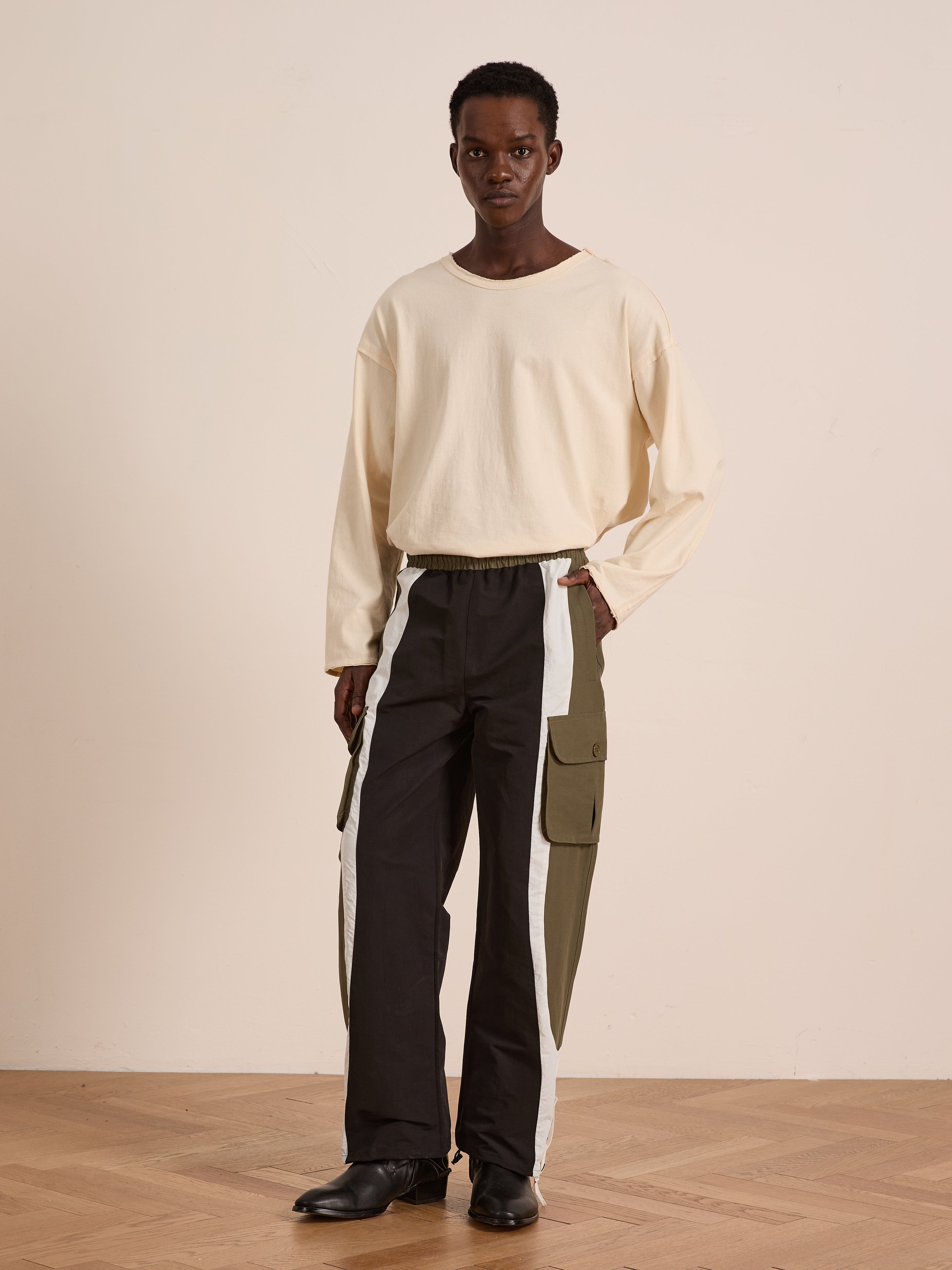 A person stands indoors wearing a long-sleeve cream shirt and Found Sporting Club Track Pants—black with olive green and white panels, adjustable waistband—paired with black shoes.