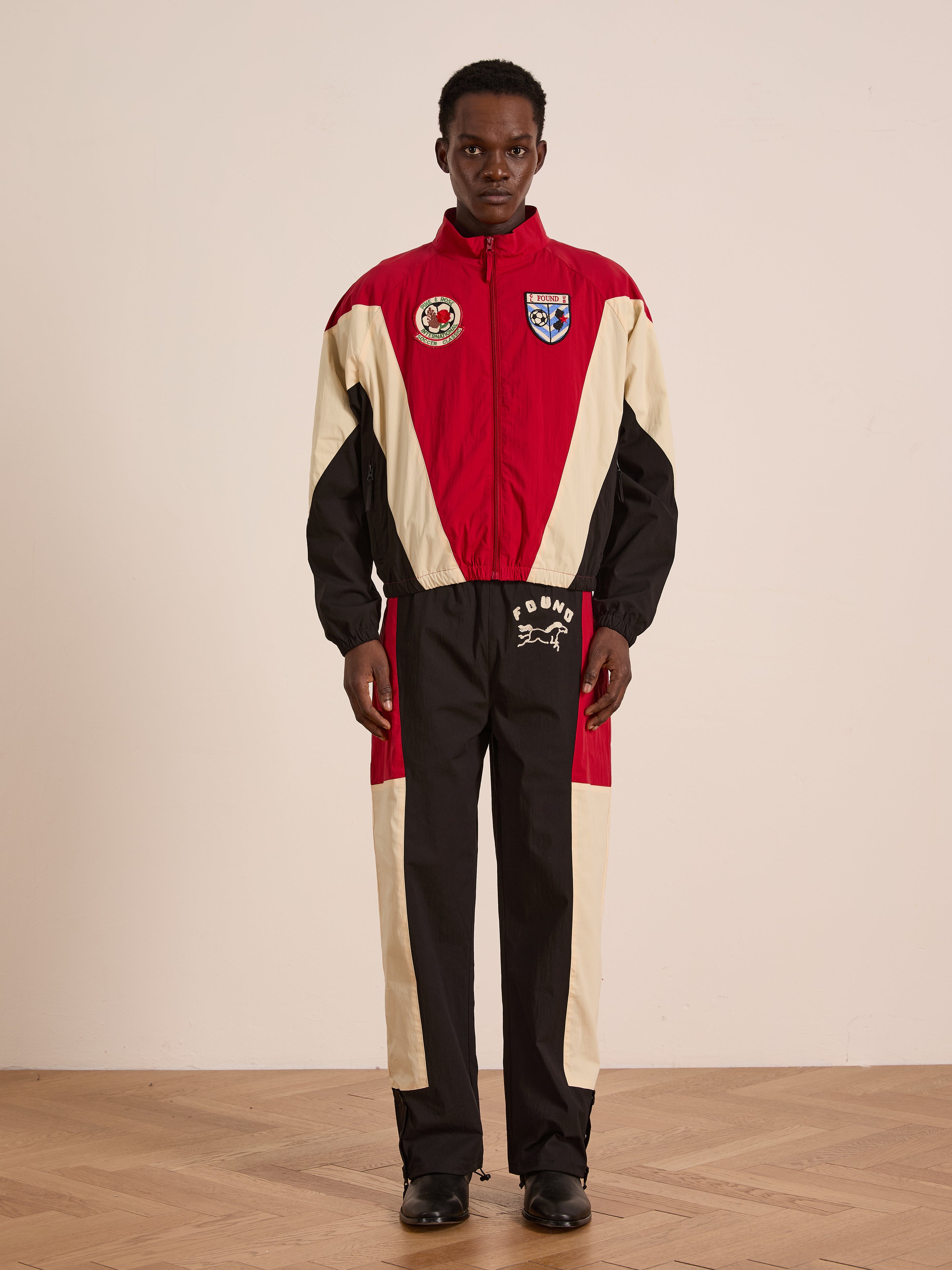 A man stands indoors wearing red, black, and beige Pine and Rose Track Pants by Found with patches and logos, black shoes, a unisex relaxed fit, and a neutral expression.