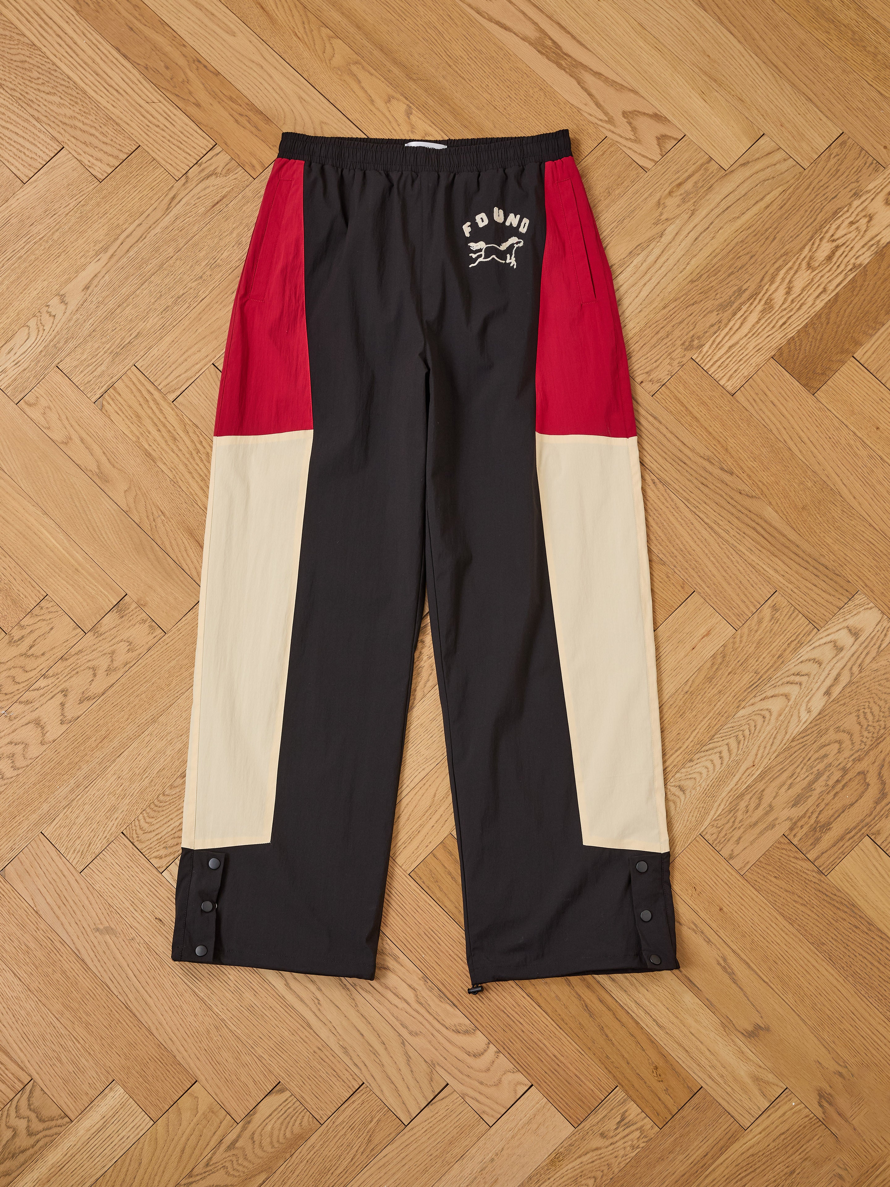 A pair of Pine and Rose Track Pants by Found, unisex relaxed fit with black, red, and beige color-block panels, elastic waistband, and "FOUND" with dog graphic on the left thigh, laid flat on a wooden floor.