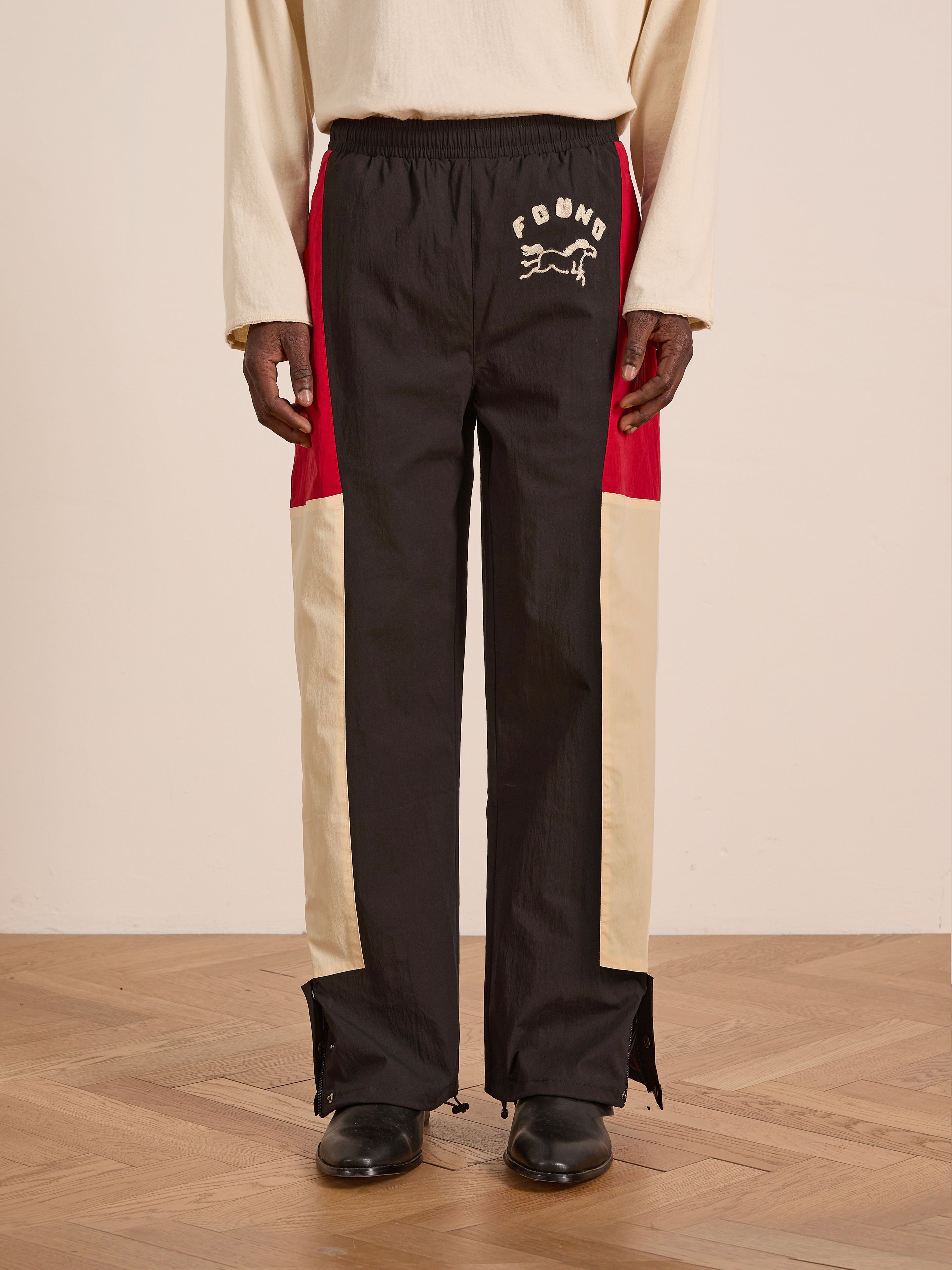 Someone stands indoors on a wood floor wearing Found's Pine and Rose Track Pants—unisex windbreaker pants in black, red, and beige with "FOUND" and a running dog graphic on the upper left thigh.
