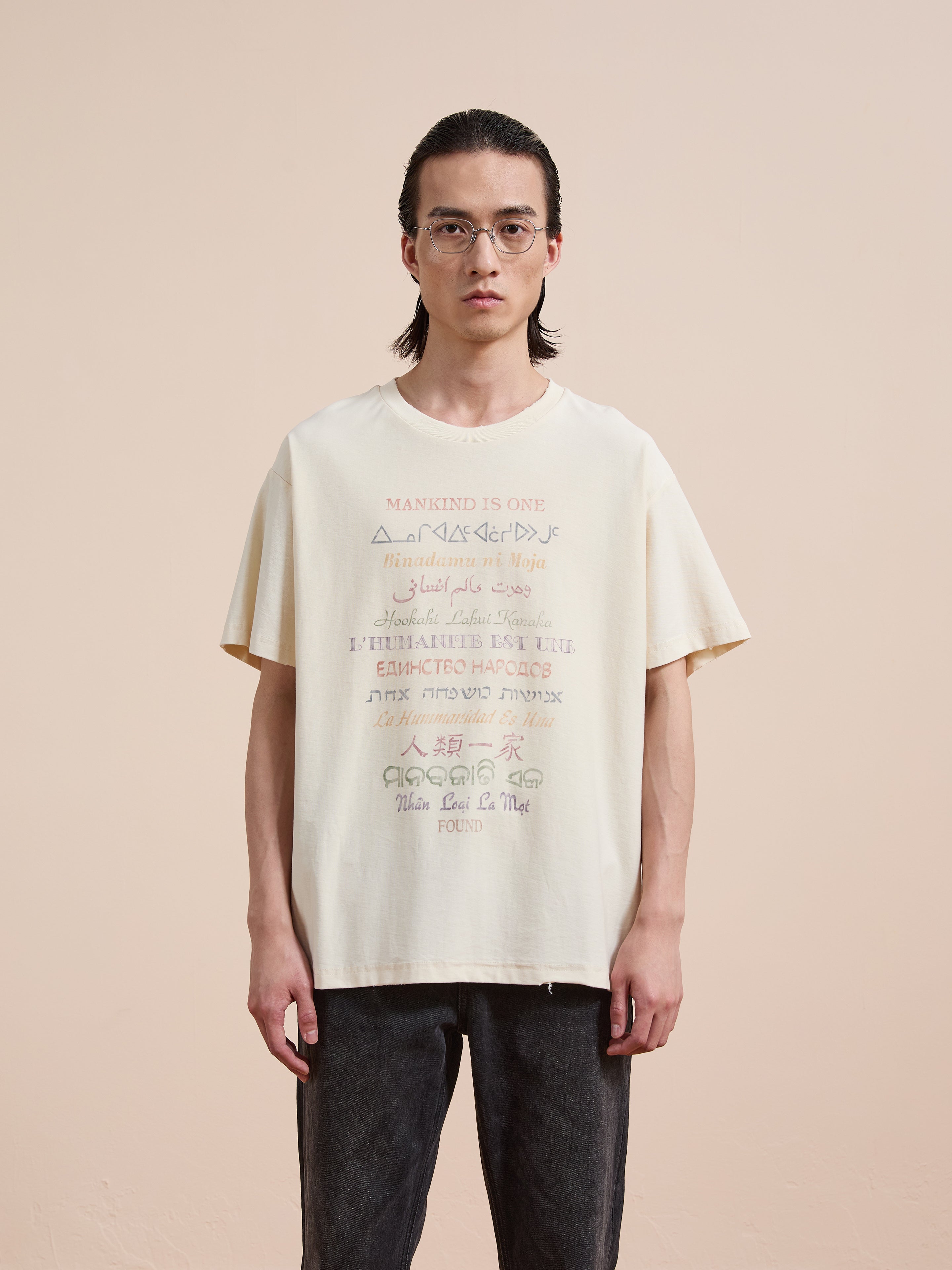 A person with long hair and glasses stands against a plain background, wearing the FOUND One Humanity Tee—a cream vintage cotton unisex t-shirt featuring multilingual text on the front—and dark pants.