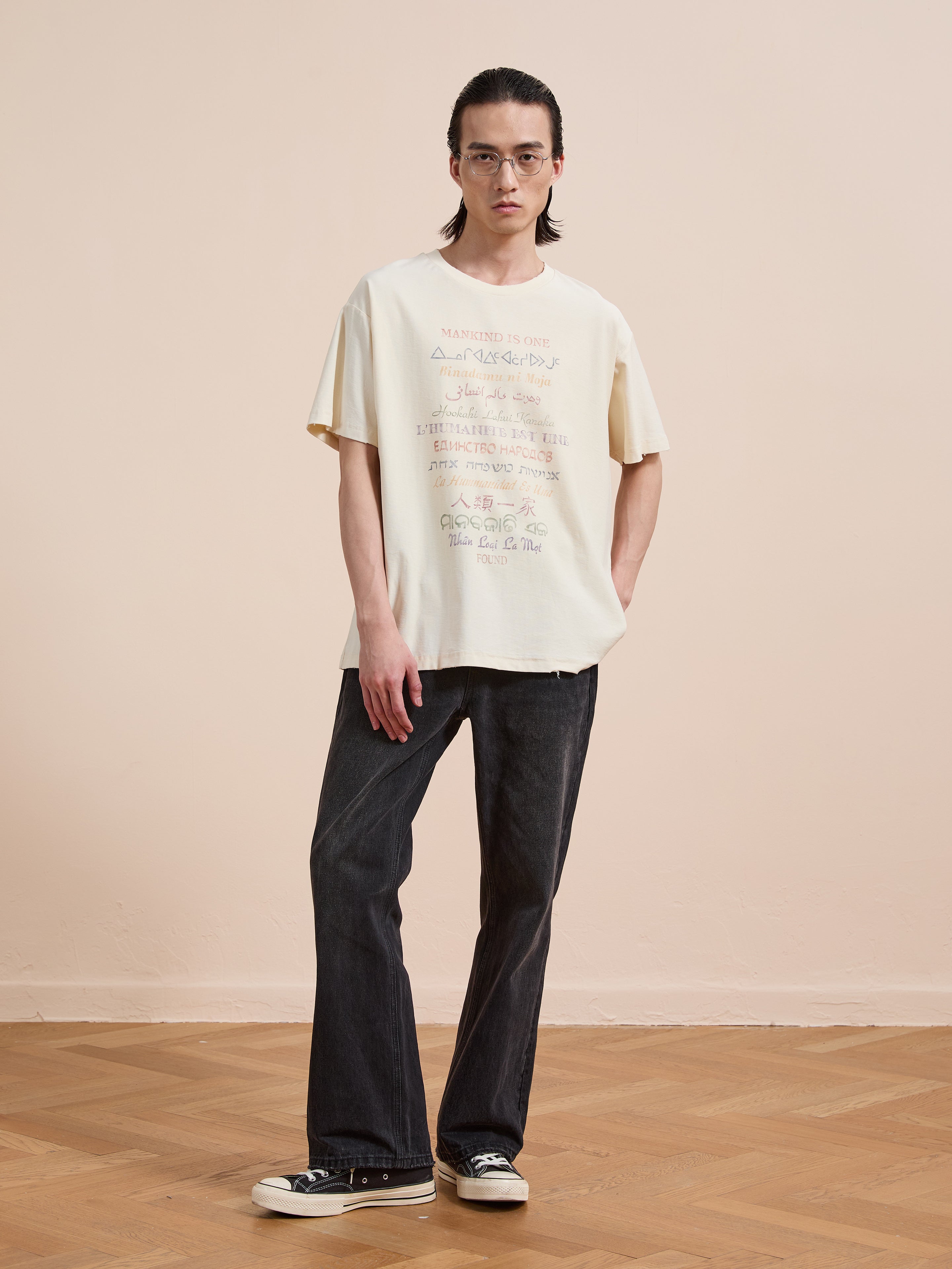A person with long hair, glasses, and a neutral expression stands indoors on a wooden floor, wearing FOUND's cream One Humanity Tee in a unisex relaxed fit, paired with black jeans and black sneakers.