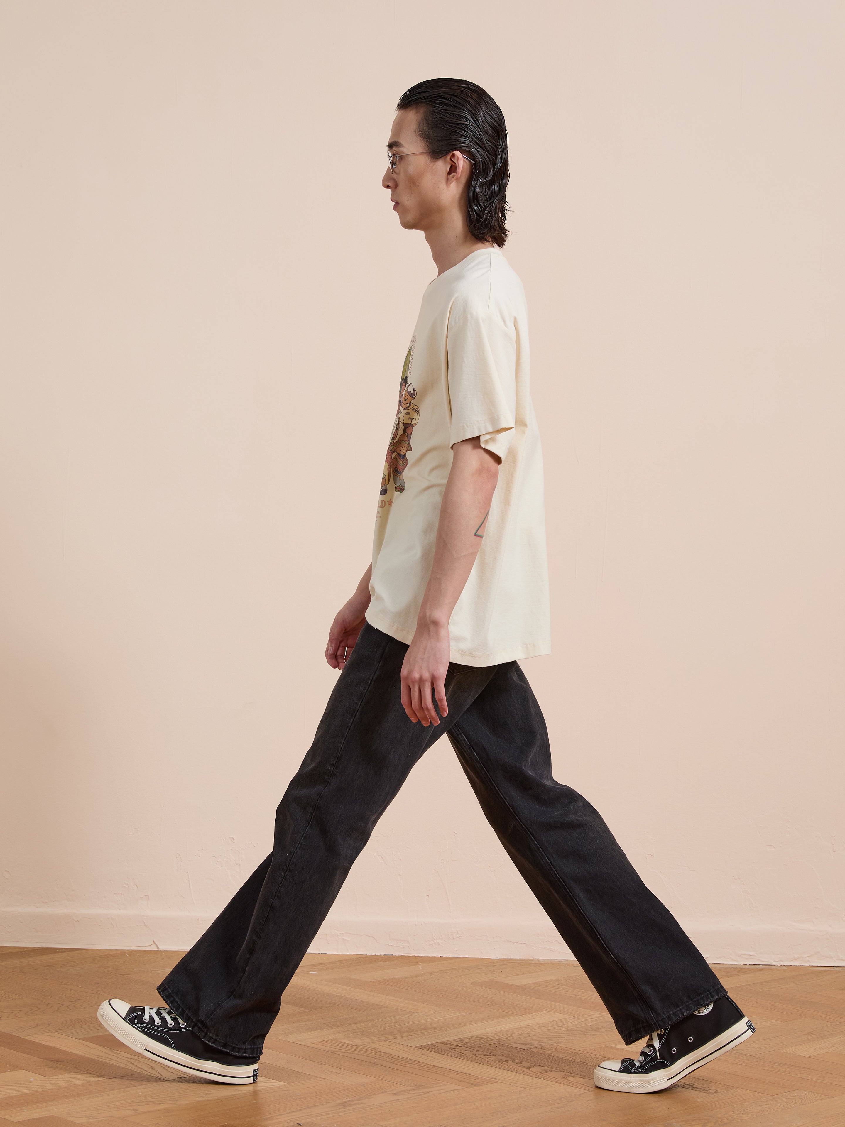 A person with long dark hair and glasses, wearing a FOUND One World Family Tee, black jeans, and black sneakers, walks indoors on a wooden floor against a plain beige wall.