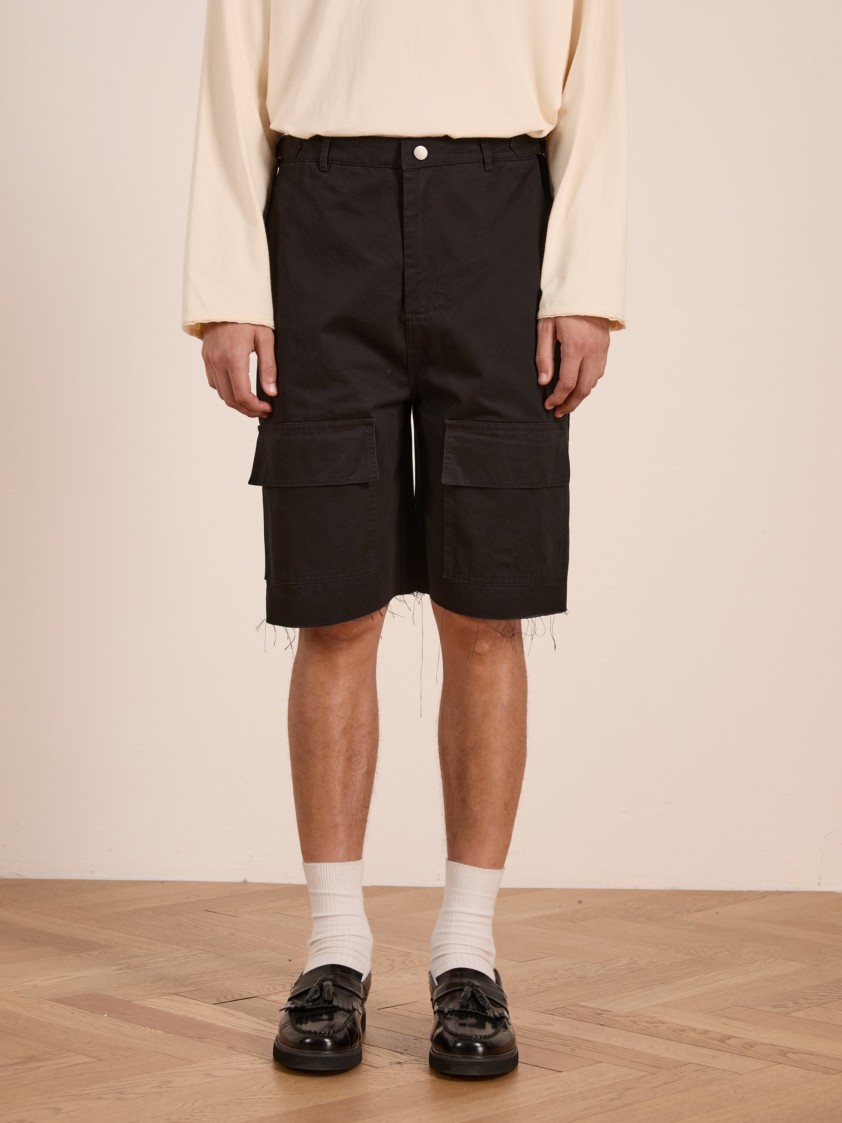 A person in a cream long-sleeve shirt, Found Parachute Cargo Twill Shorts in black, white socks, and black loafers stands on a wooden floor against a plain light wall, showcasing modern utility wear.