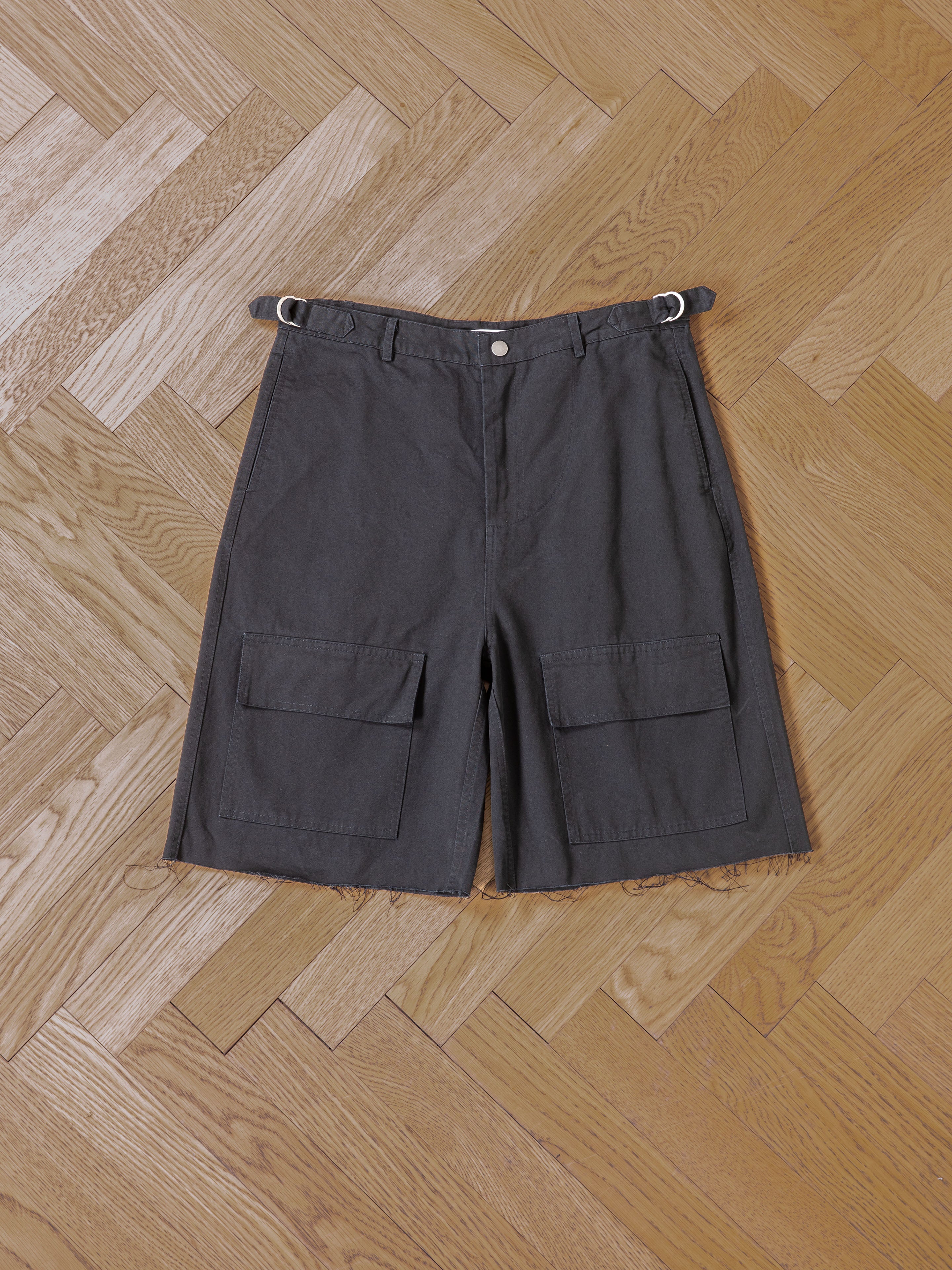 Found Parachute Cargo Twill Shorts in dark gray, featuring front flap pockets and metal buckle details, shown laid flat on a patterned wooden floor—perfect for modern utility style.