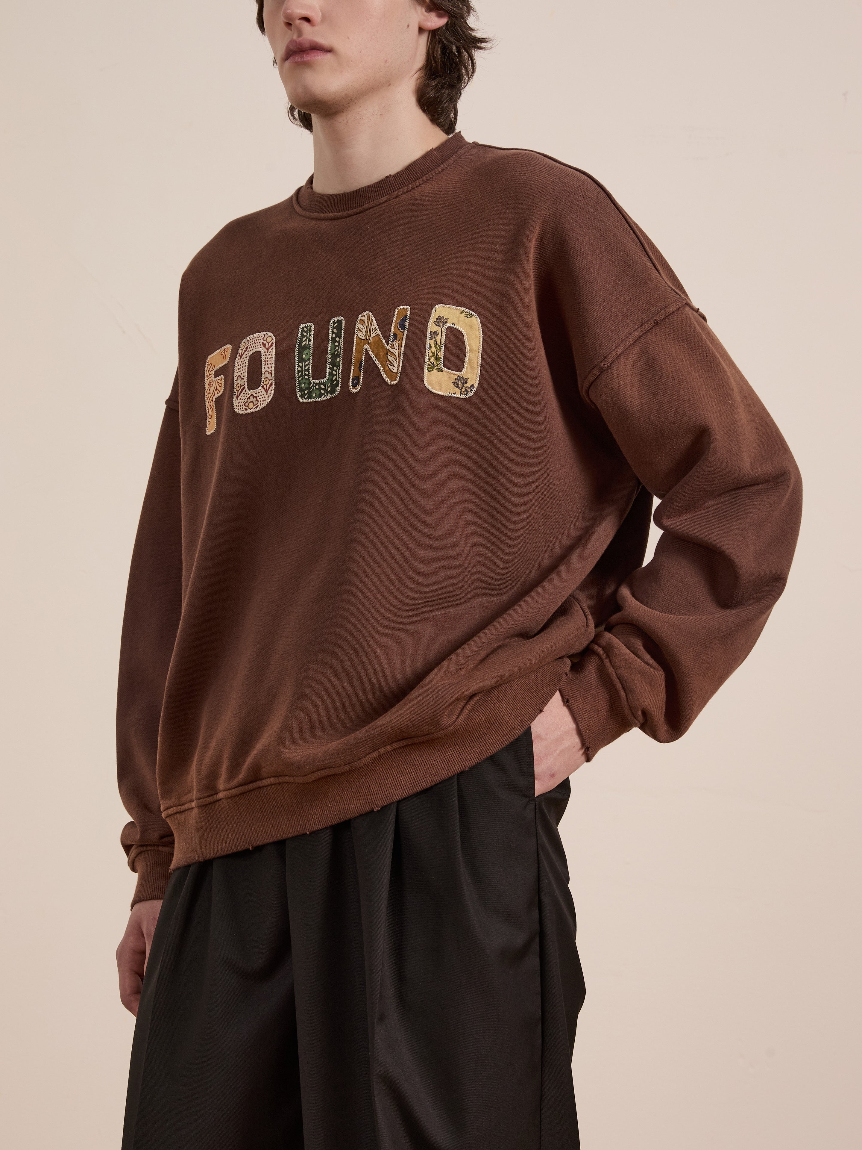 A person wearing a Patterned Logo Crewneck from FOUND, featuring brown patterns and appliqué "FOUND" lettering, paired with black pants.