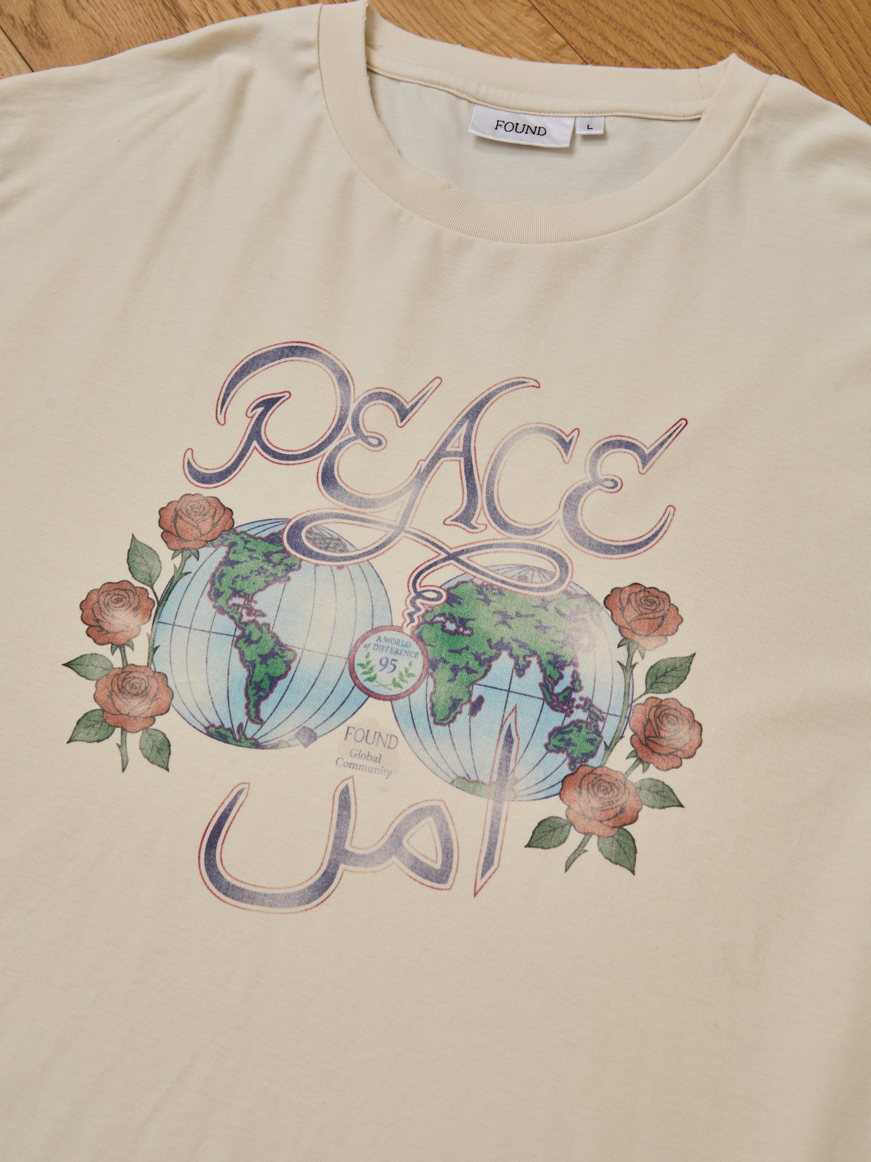 The FOUND Peace Globe Tee is a unisex, relaxed fit vintage cotton t-shirt in beige, featuring "PEACE" above two globes, roses, and "peace" in Arabic script. The tee is laid flat on a wooden surface.