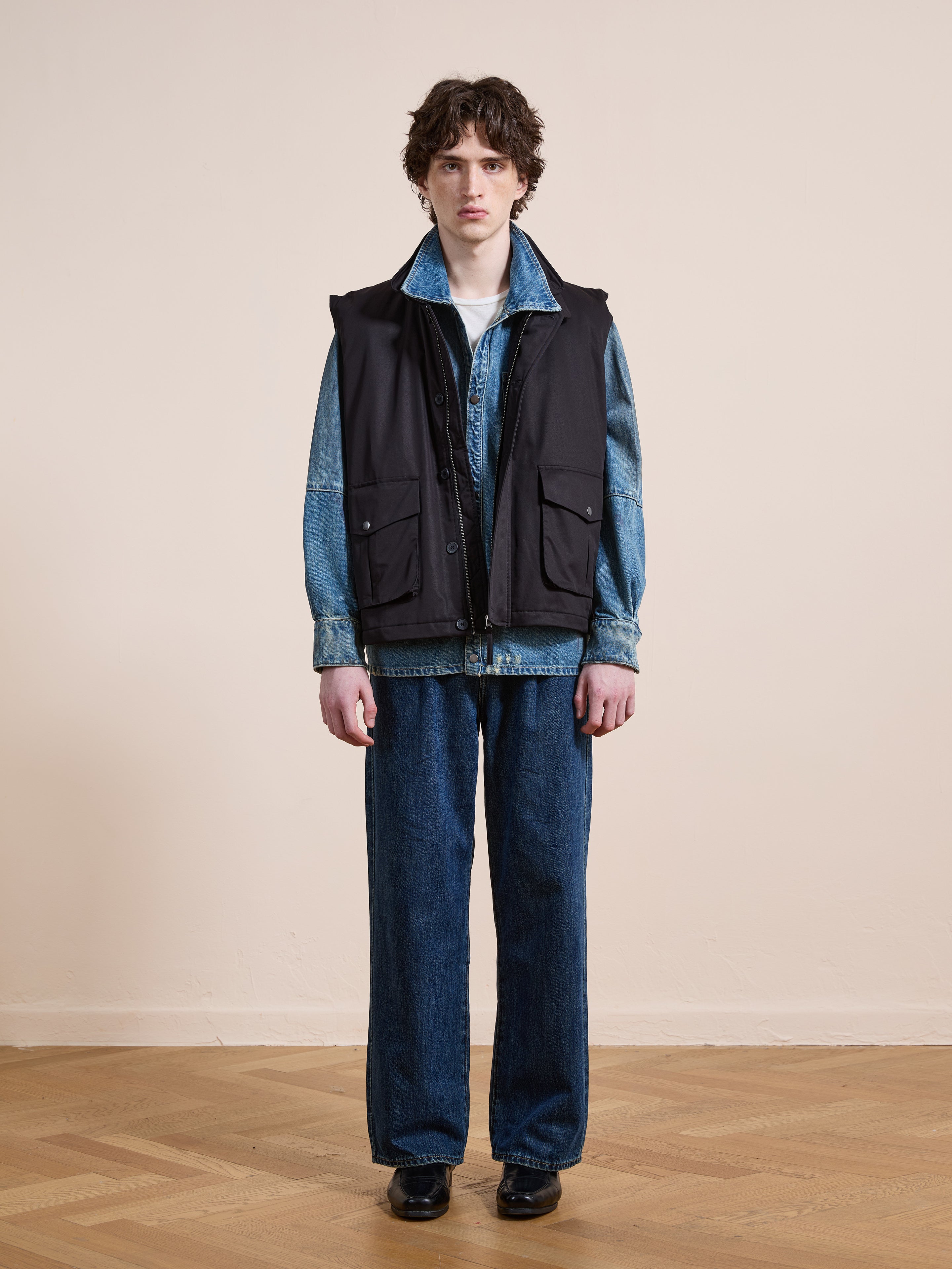 Someone stands indoors against a beige wall and wooden floor, wearing FOUND's Padded Cargo Vest over a denim jacket with blue jeans and black shoes.