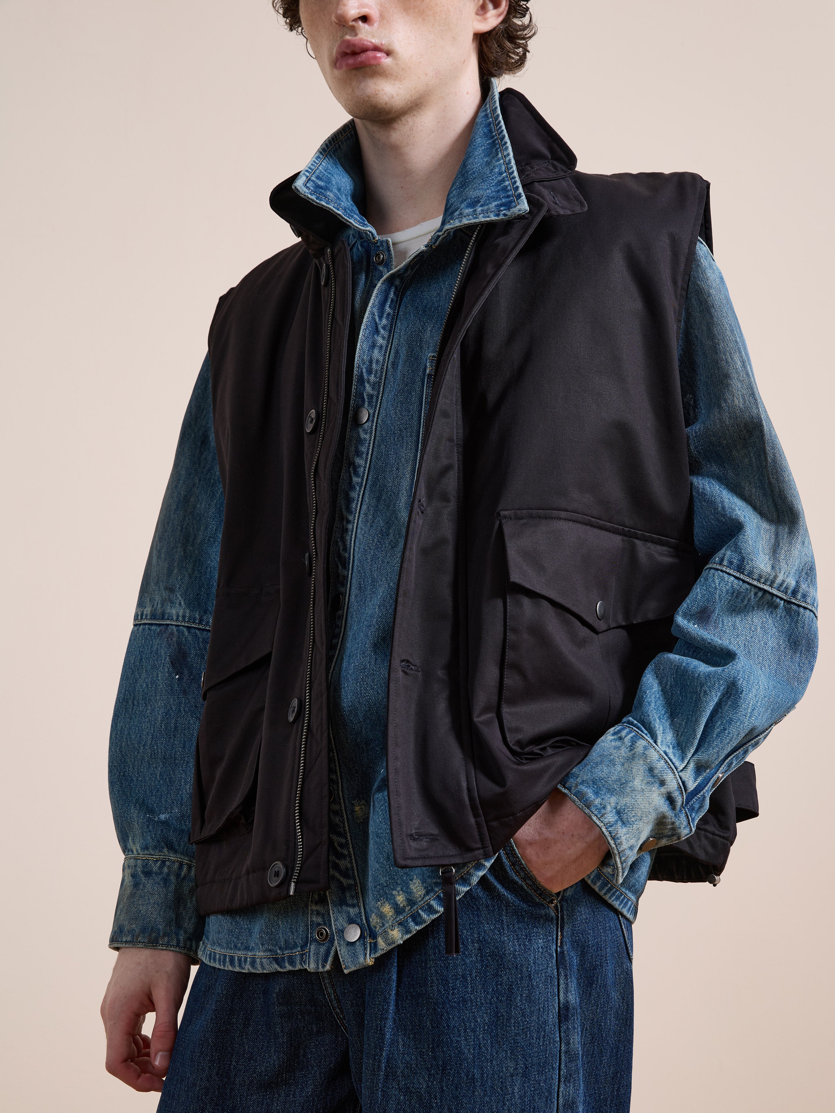 A person wears the FOUND Padded Cargo Vest in black over a blue denim jacket and jeans, standing with one hand in their pocket against a plain background.