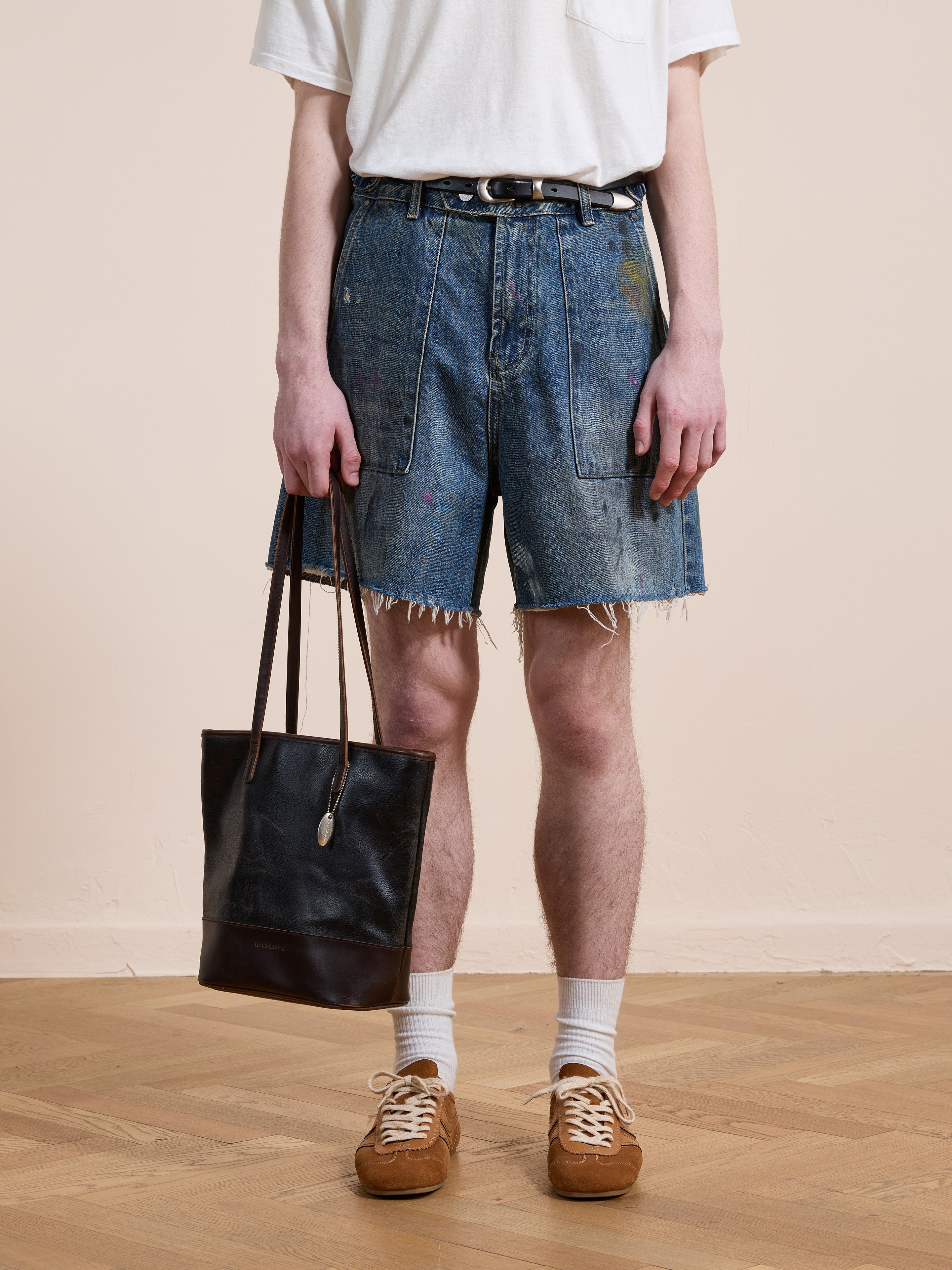 A person in a white t-shirt, Found Paint Studio Denim Shorts, brown sneakers with white socks, and holding a black leather tote bag stands on a wooden floor.