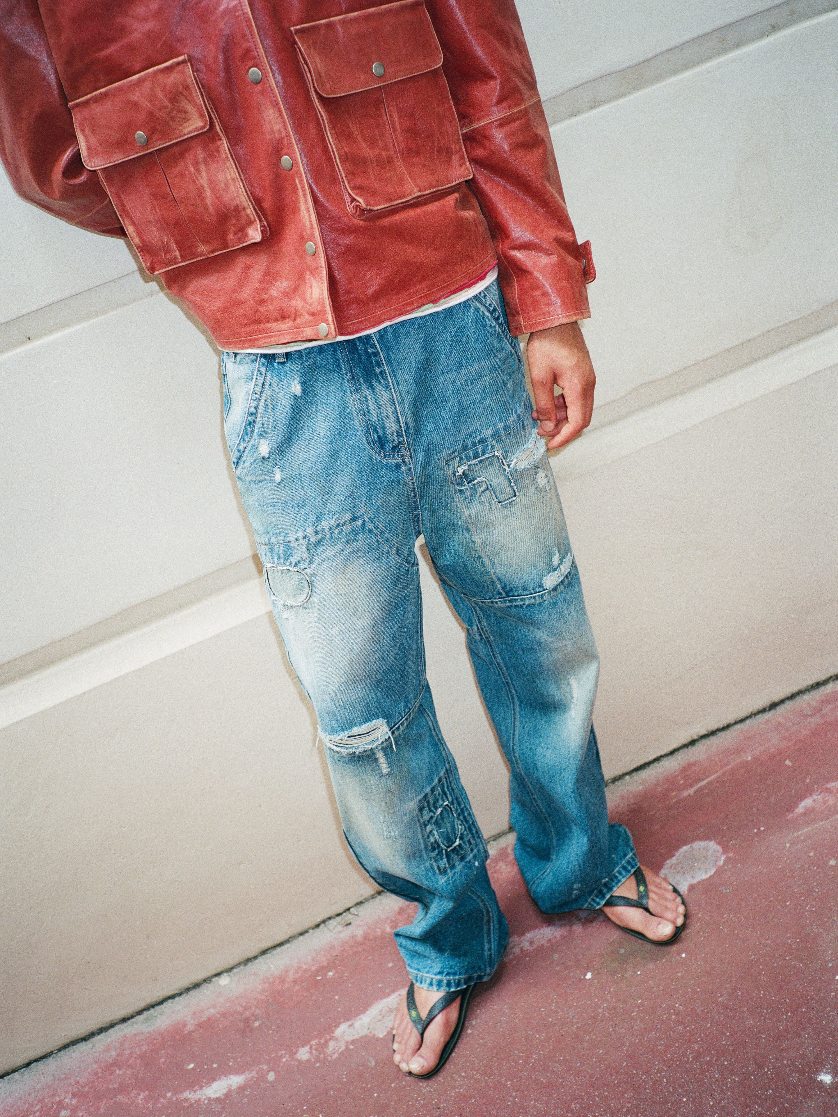 A person wearing FOUND Painter Repaired Jeans, a red leather jacket, and black flip-flops stands against a light-colored wall.