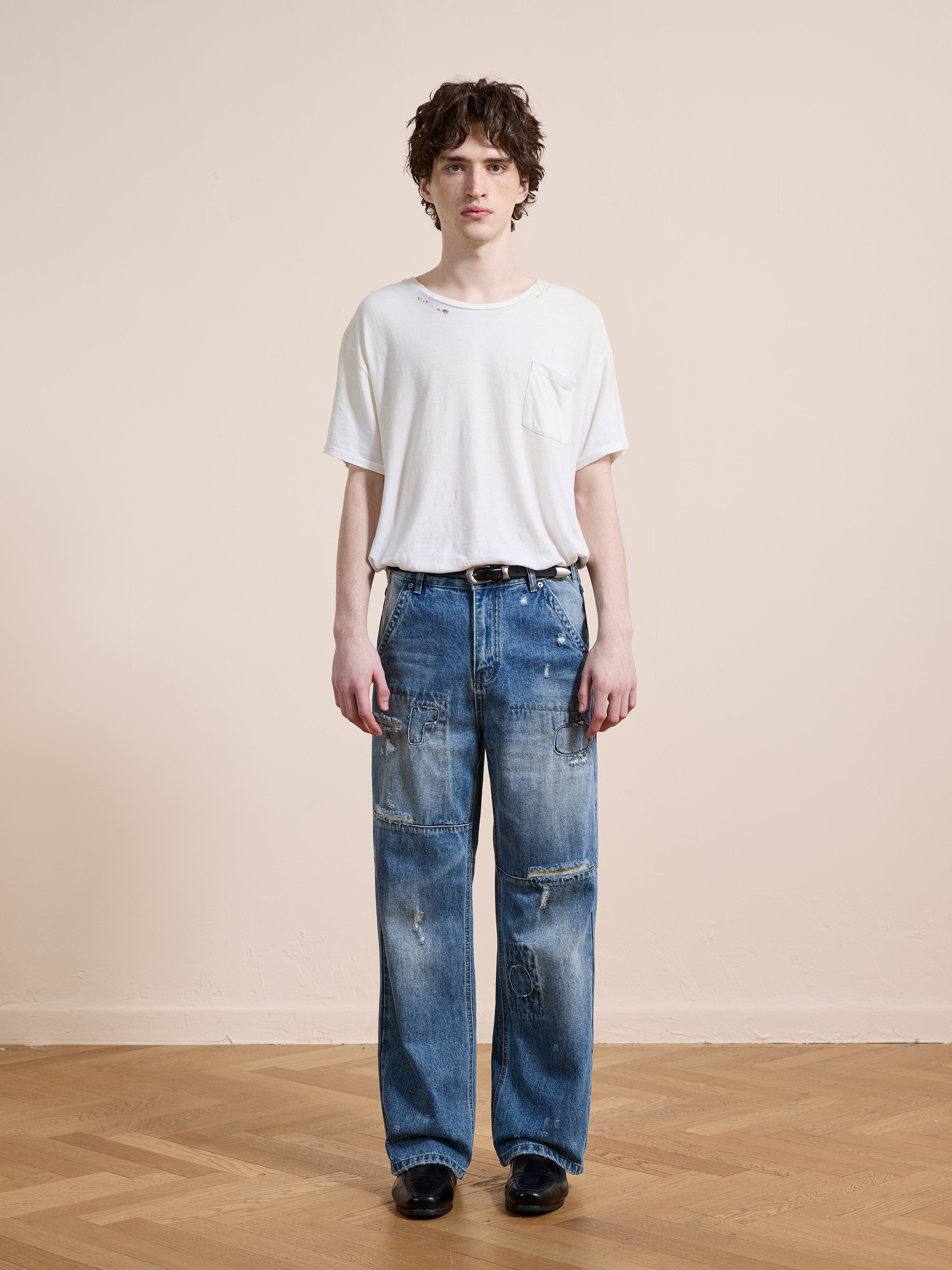 A person stands indoors on a wooden floor, facing the camera in FOUND Painter Repaired Jeans and a relaxed fit white t-shirt with holes, wearing a neutral expression.