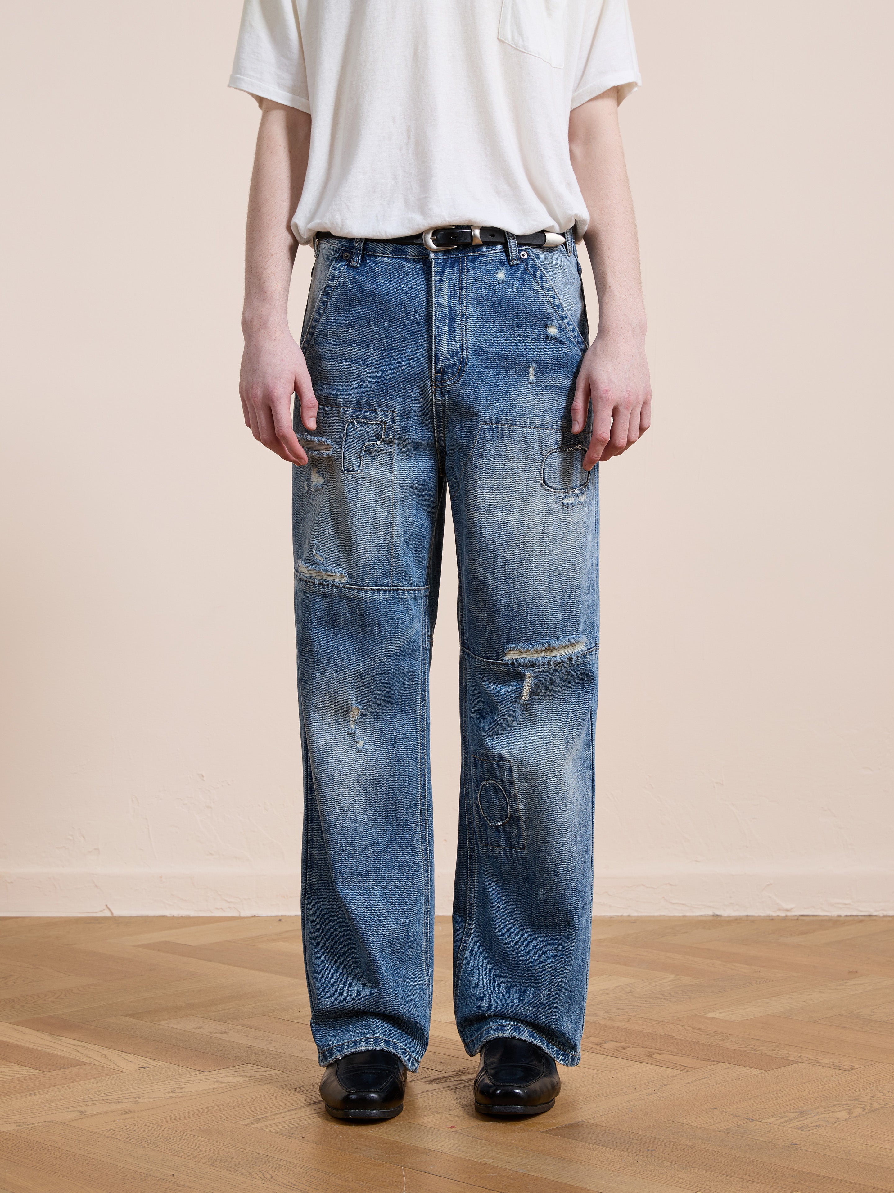 A person wearing a white t-shirt, FOUND Painter Repaired Jeans featuring vintage denim with patches and rips, paired with a black belt and black shoes, stands on a wooden floor against a plain light-colored wall.