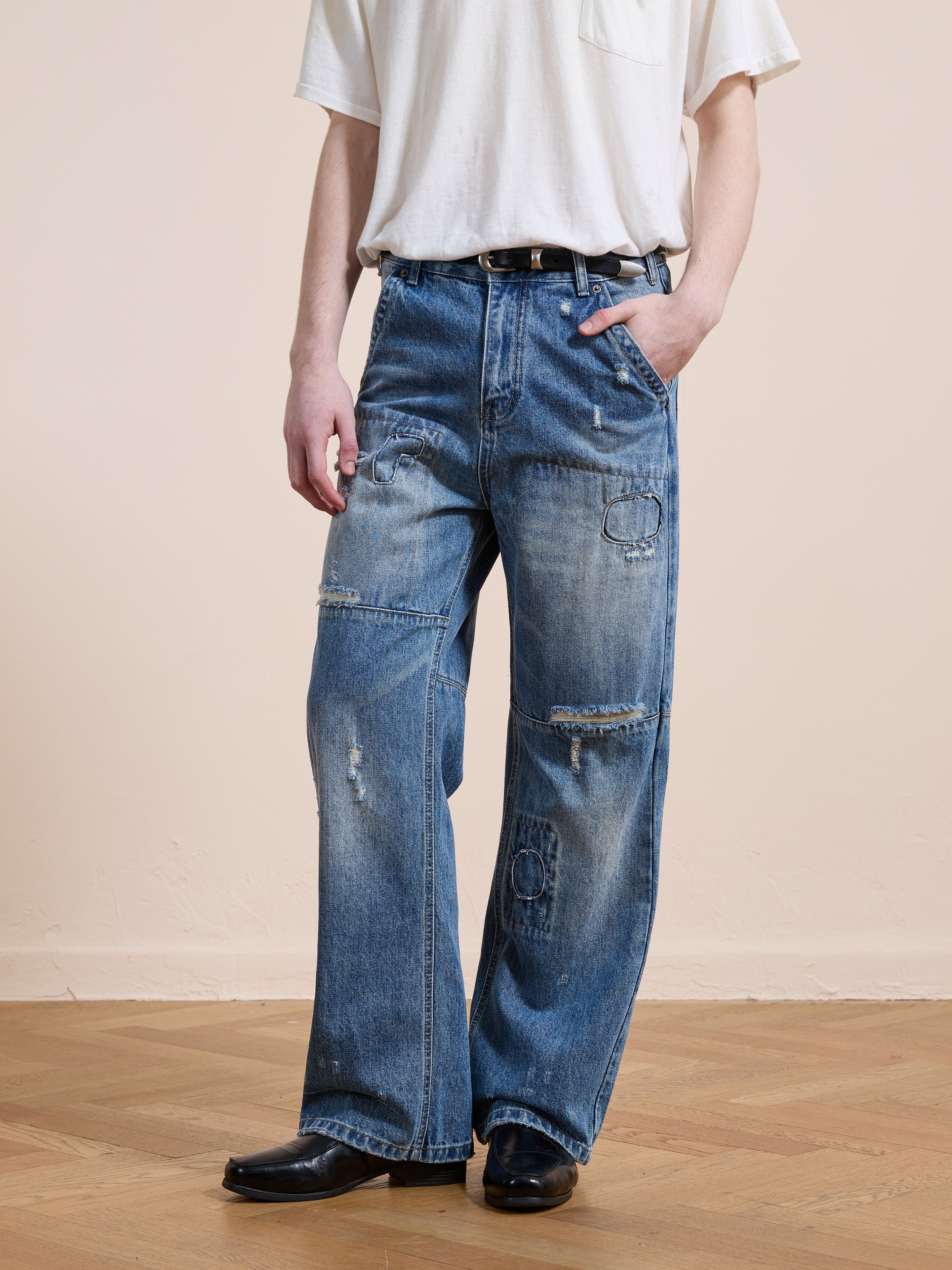 A person wears FOUND Painter Repaired Jeans—vintage denim with distressed, patchwork details—paired with a white t-shirt and black shoes, standing on a wooden floor against a plain background.