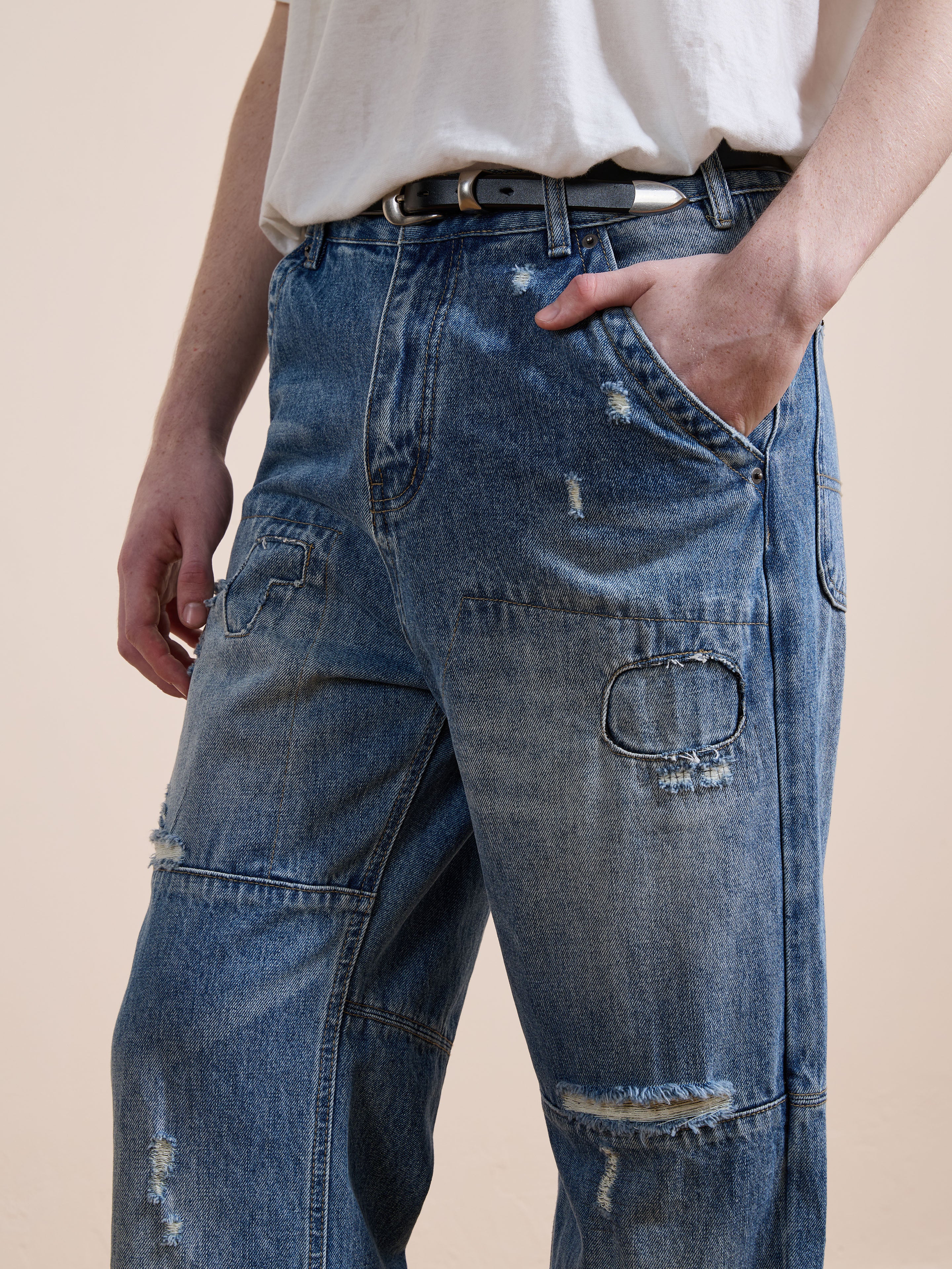 A person wearing FOUND Painter Repaired Jeans in vintage denim with a relaxed unisex fit, a white t-shirt, and black belt, stands hands-in-pockets against a plain beige background.