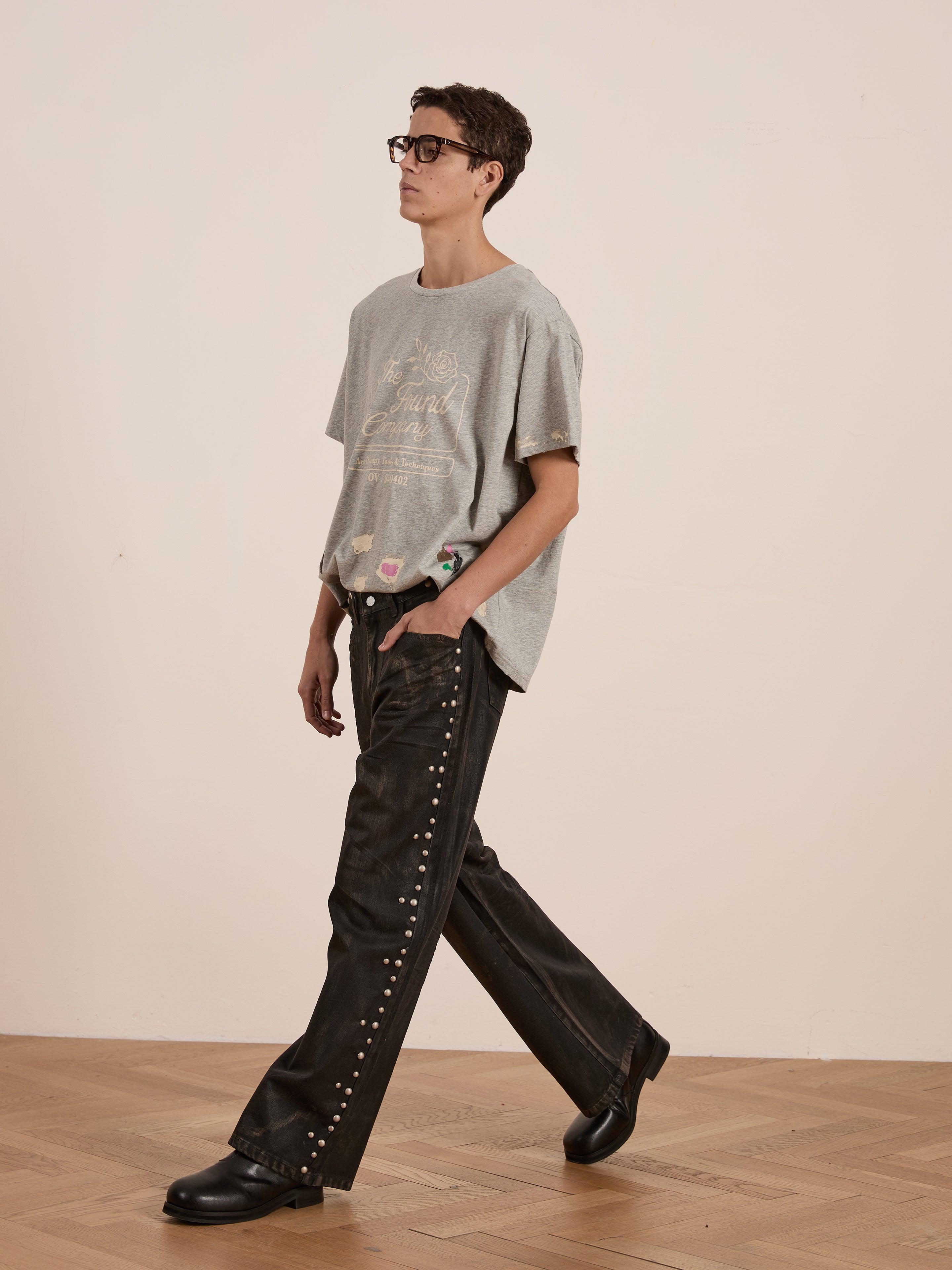 Sporting the Painters Company Tee by FOUND and black studded pants, someone captures a workwear-inspired look on a wooden floor. Their outfit is completed with unisex black shoes and vintage-style glasses, all set against a plain backdrop.