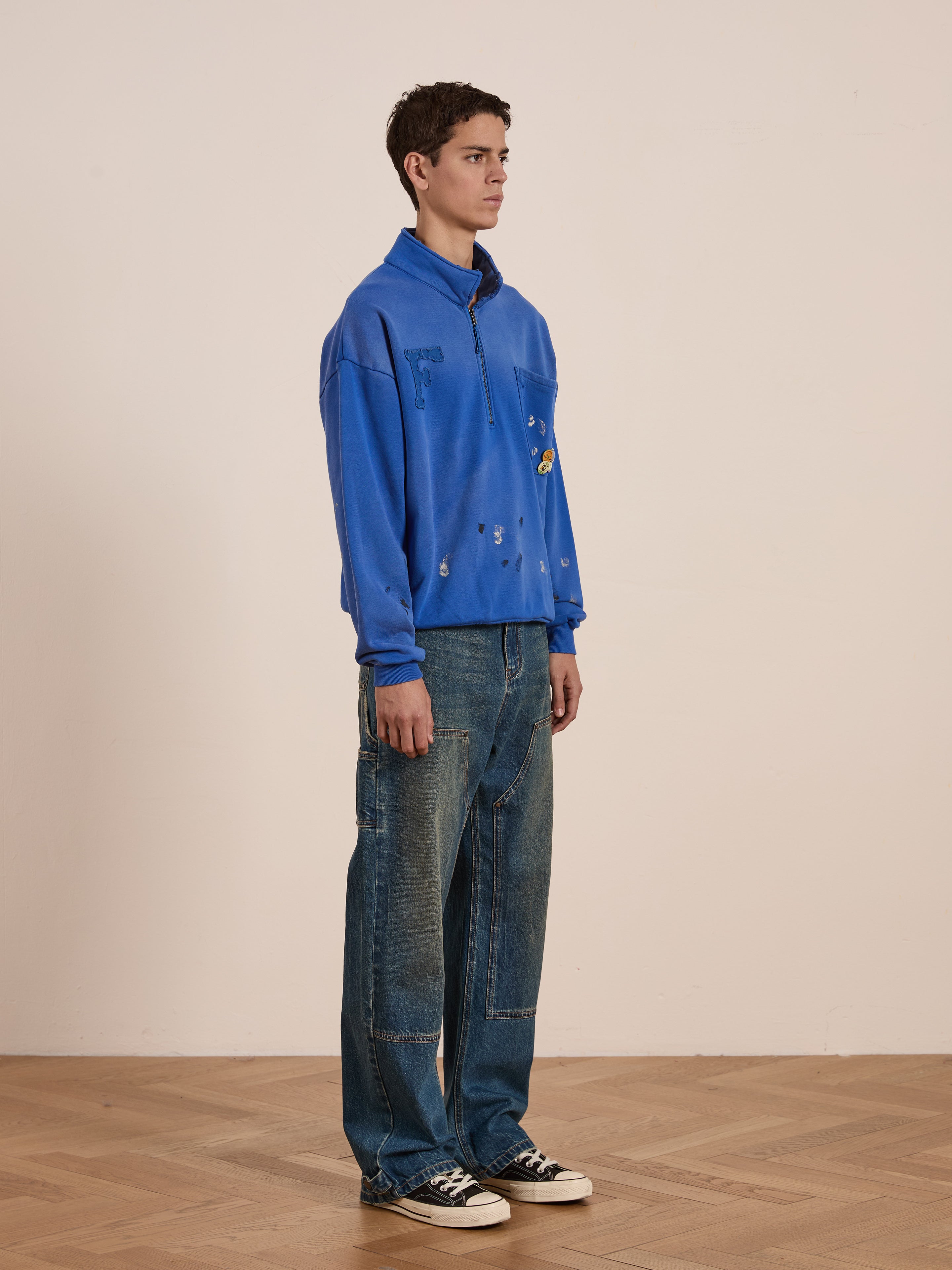 A person stands on a wooden floor, wearing a blue zip-up jacket with small designs, FOUND vintage-inspired Denim Carpenter Jeans, and black sneakers, facing slightly to the left against a plain background.