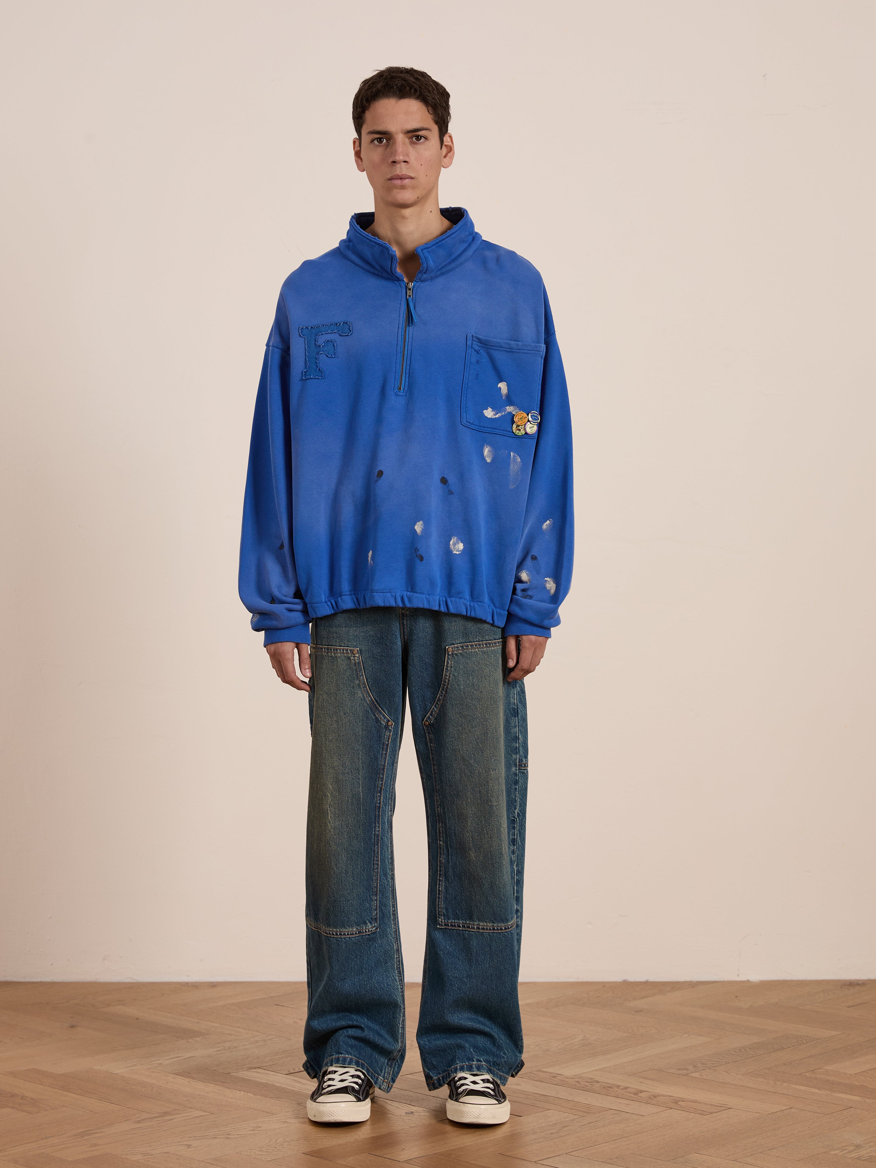A person in a blue hoodie featuring decorative elements and wearing FOUND's Denim Carpenter Jeans, known for their vintage-inspired, relaxed straight-leg fit, stands on a wooden floor against a plain wall.