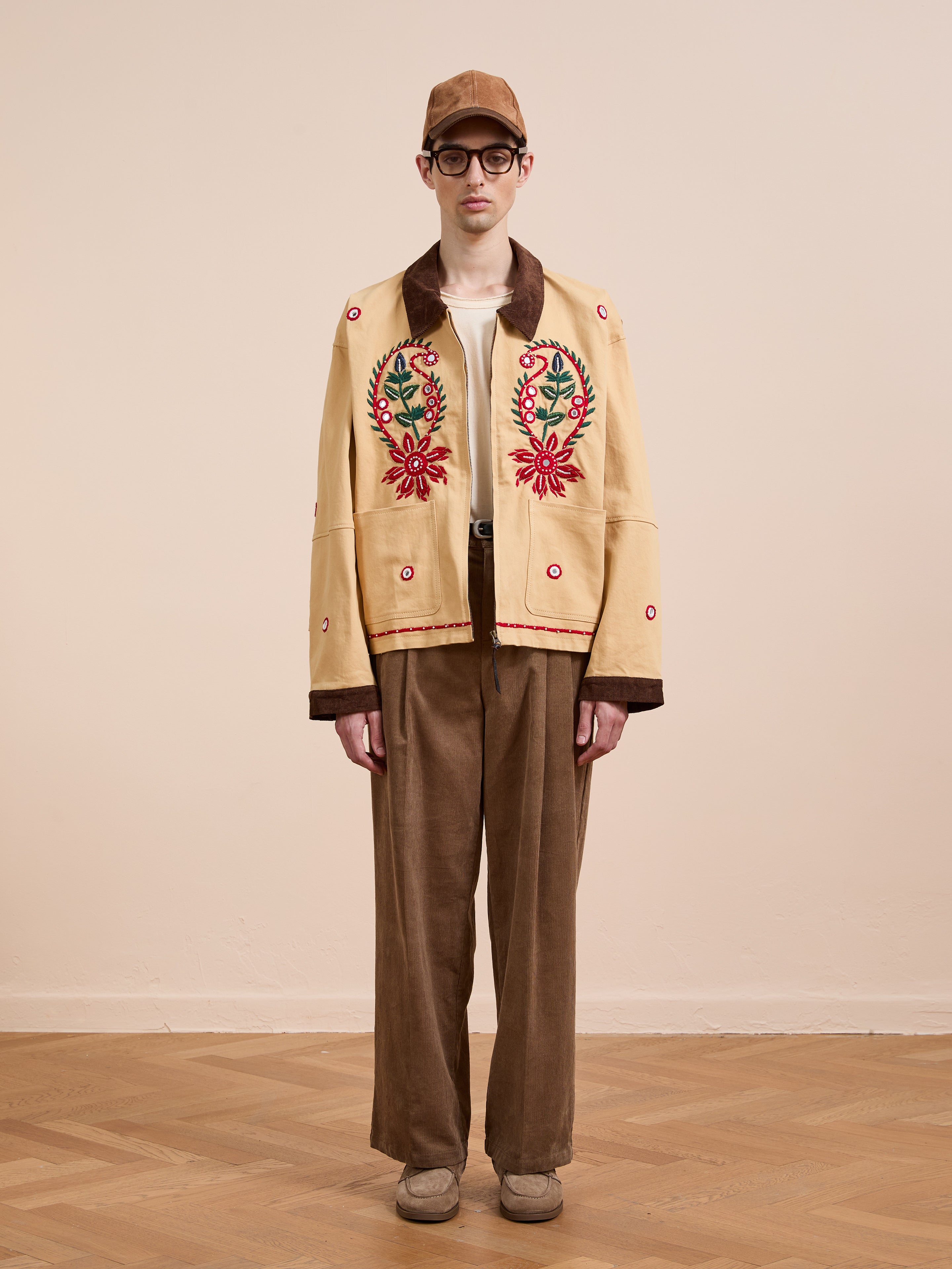 A person stands indoors against a neutral background wearing the Found Paisley Motif Hand Embroidered Work Jacket in tan, paired with brown pants, a brown cap, glasses, and beige shoes.