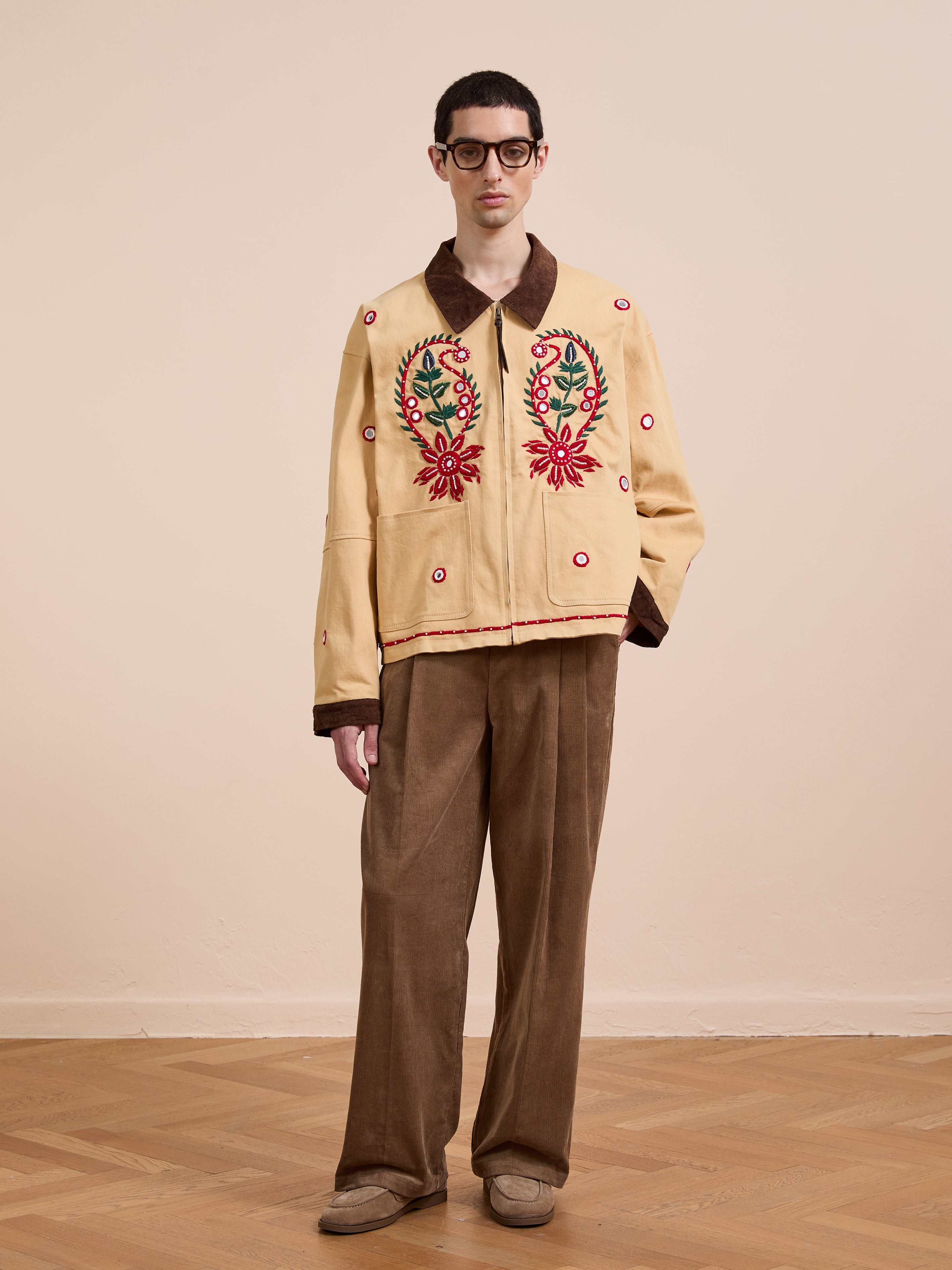 A man stands indoors wearing glasses, brown pants, beige shoes, and the Found Paisley Motif Hand Embroidered Work Jacket with colorful paisley floral embroidery and a brown collar, set against a plain background.