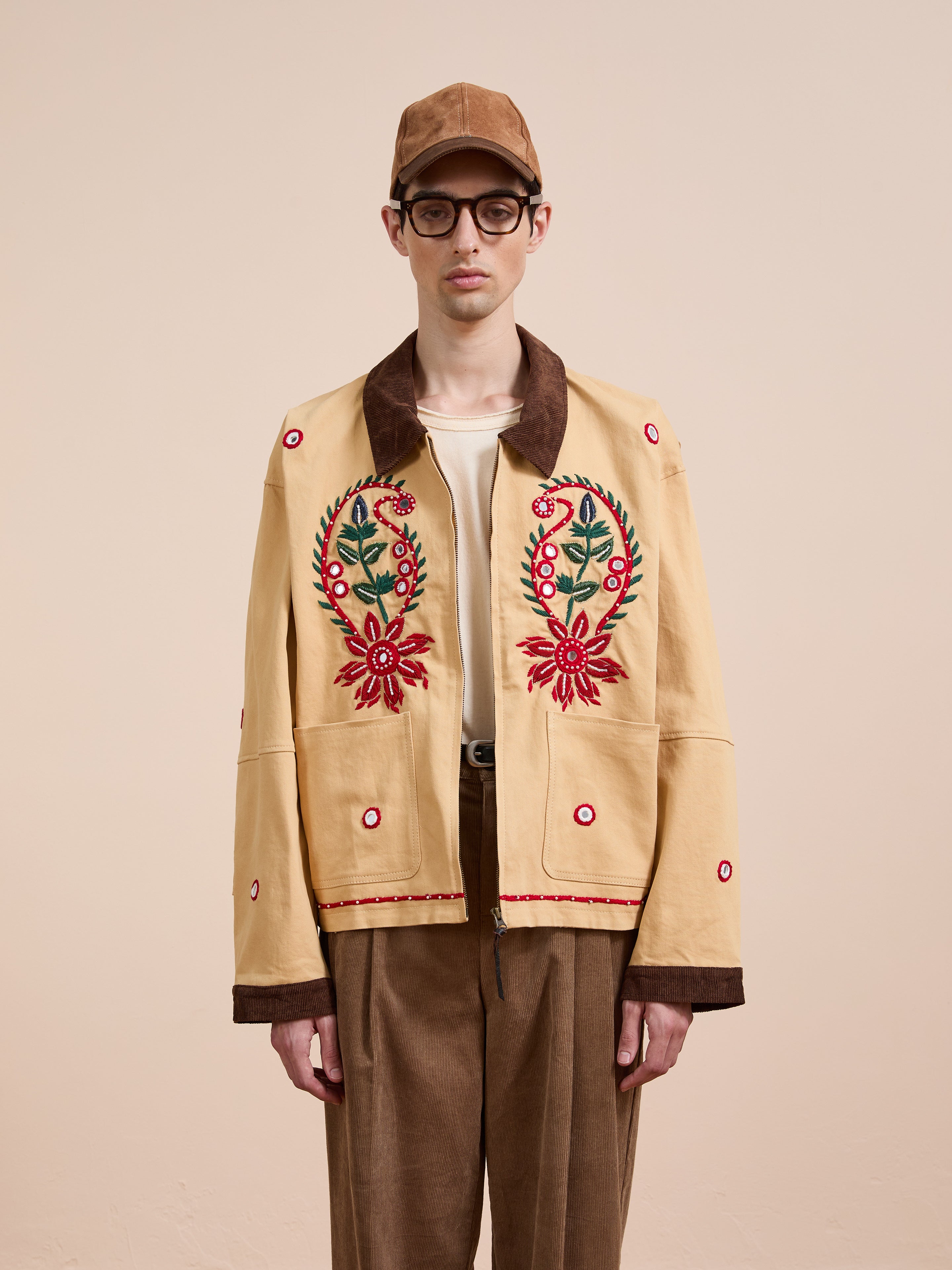 Someone wearing the Found Paisley Motif Hand Embroidered Work Jacket, brown cap, glasses, cream shirt, and brown pants stands against a plain beige background.